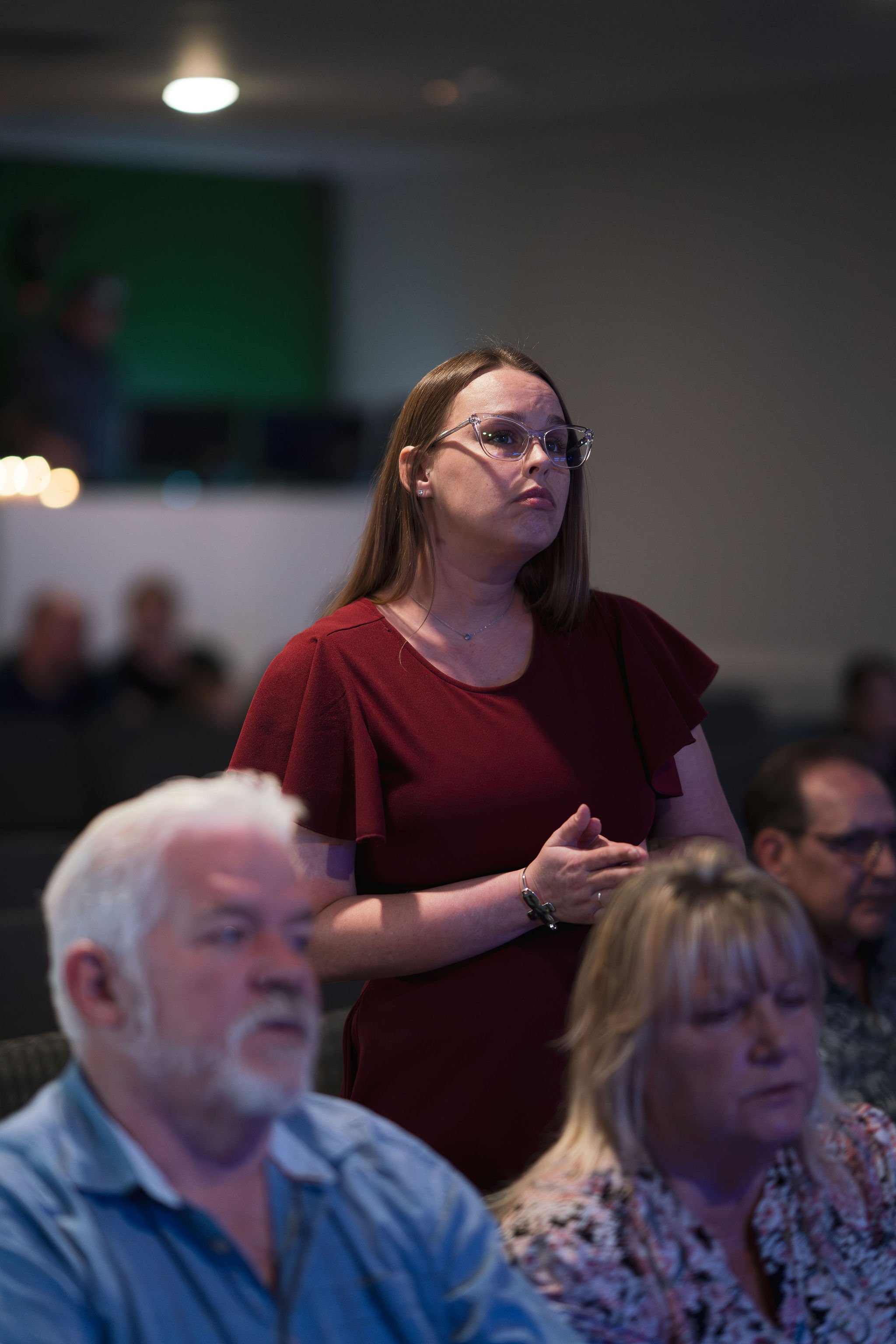 A woman standing and speaking at a conference or event, wearing glasses and a red blouse, with an audience seated around her.