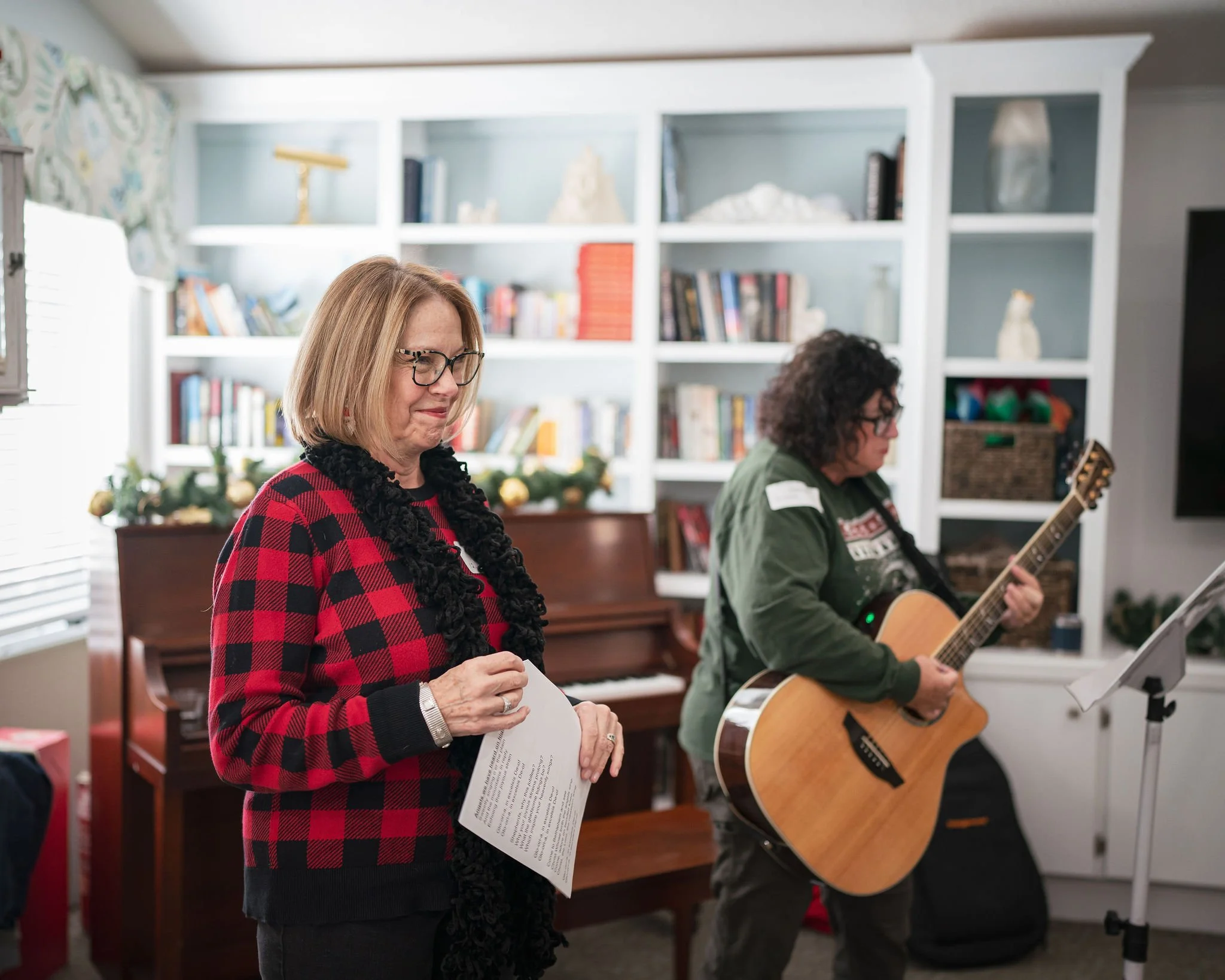 Two women are in a room decorated with a white bookshelf, playing music and singing. One woman is holding sheet music, wearing glasses and a red and black checkered sweater. The other woman is playing an acoustic guitar, wearing glasses and a green j
