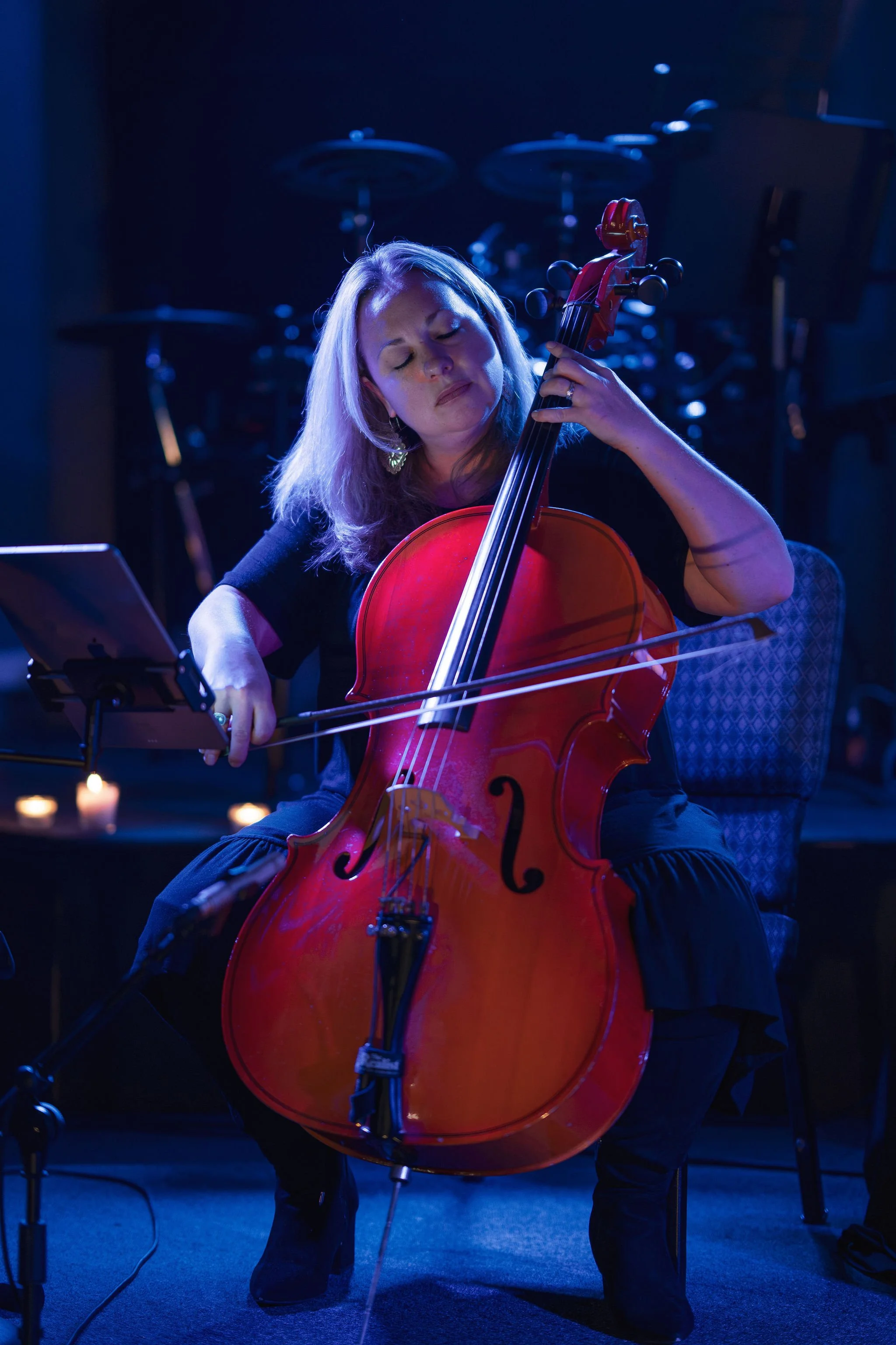 A woman playing a red cello during a live performance, with sheet music on a stand and microphones around her, in a dimly lit setting with blue lighting.