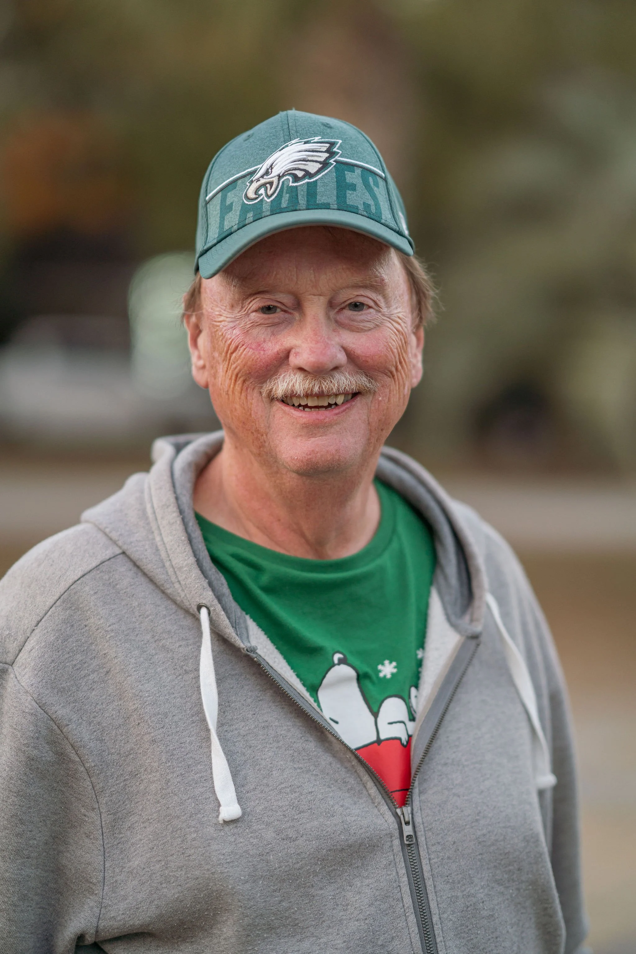 A smiling man with light skin, pale blue eyes, and a mustache, wearing a green Philadelphia Eagles cap and a gray zip-up hoodie over a green Christmas sweater with a snowman design, outdoors with blurred background.
