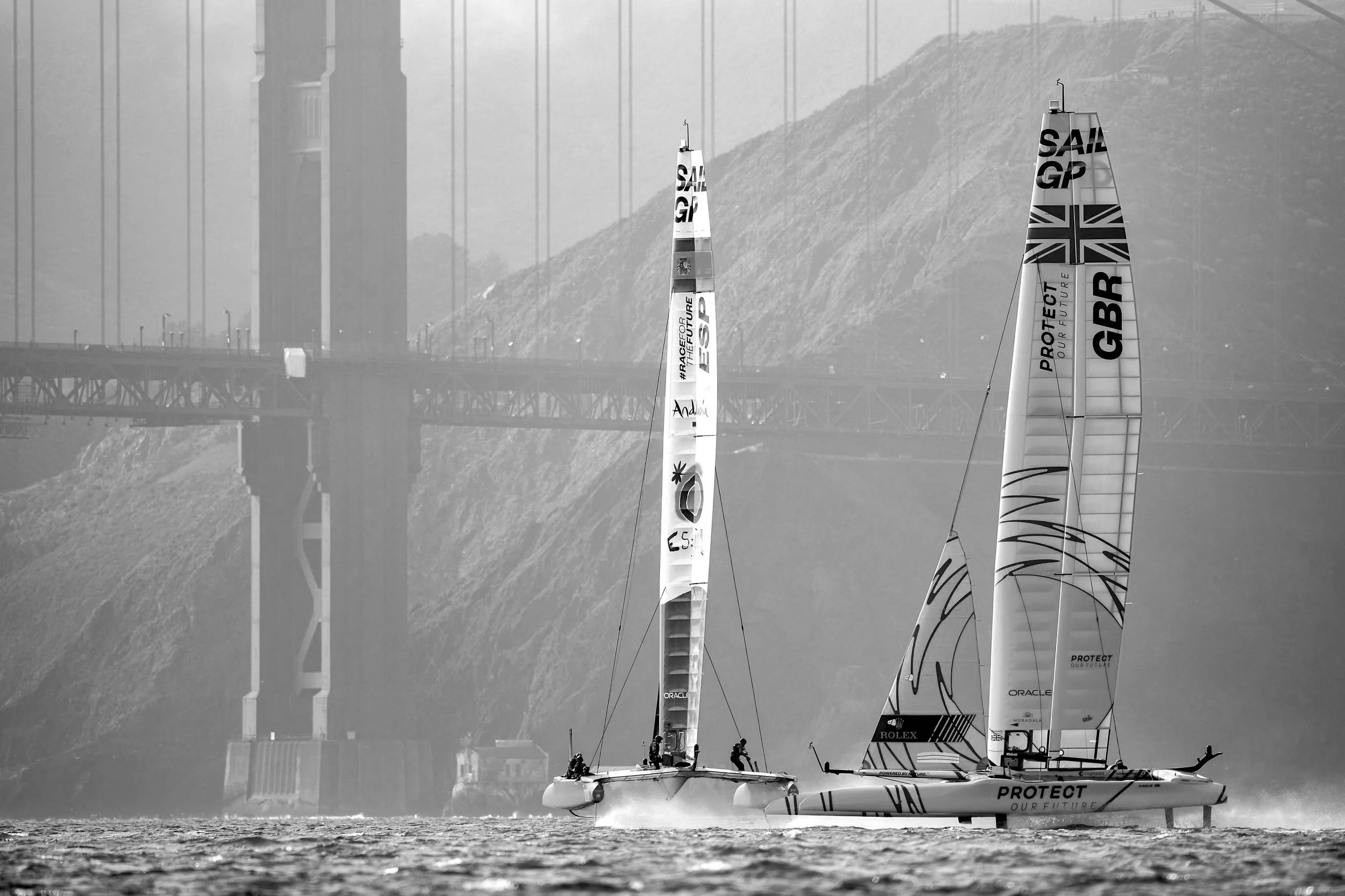 Black and white photo of two sailboats racing on water, with their sails displaying sponsorship branding, and a bridge and hilly terrain in the background.