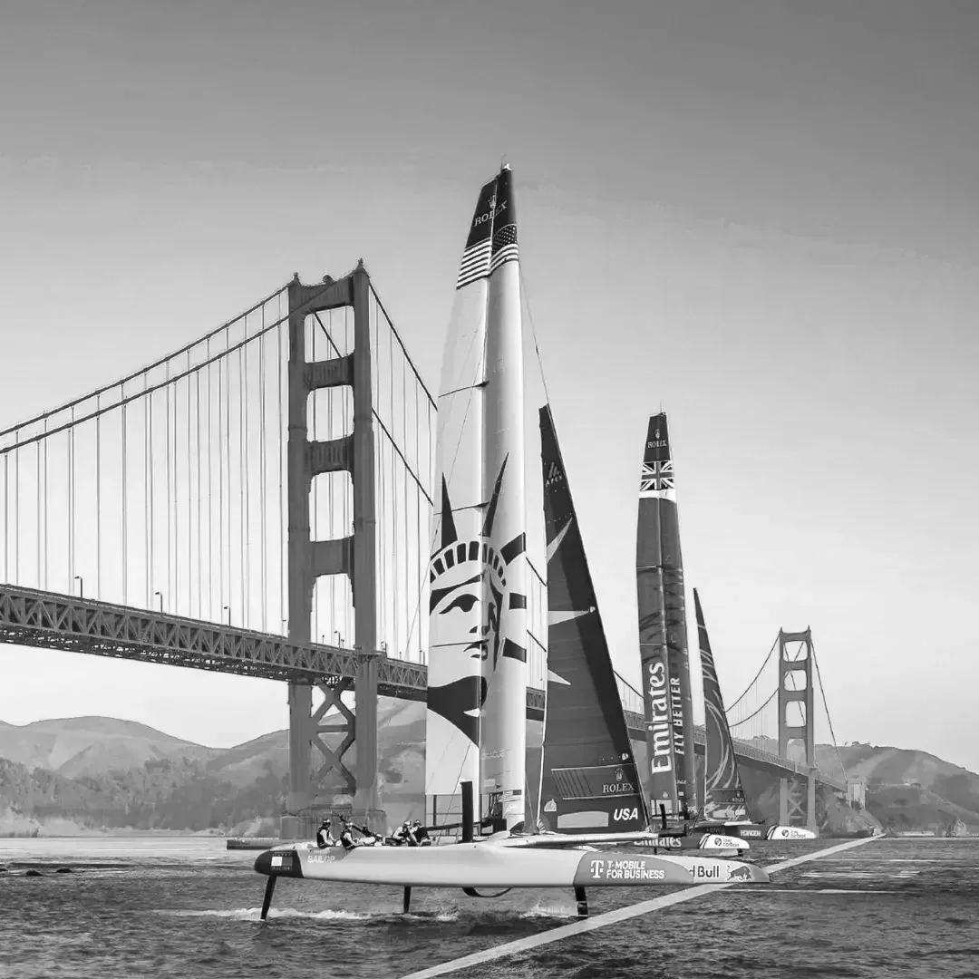 Three futuristic sailboats race near the Golden Gate Bridge on the water, with hills in the background, in black and white