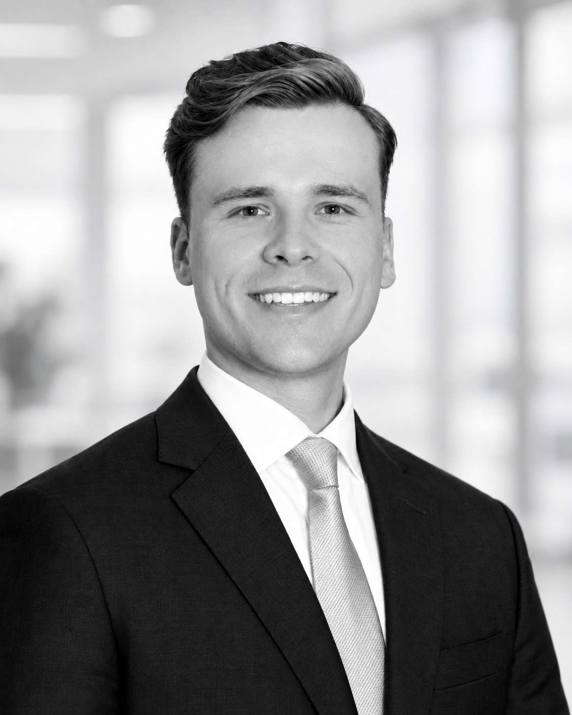 Black-and-white headshot of a young man wearing a suit, white shirt, and tie, smiling, with a blurred office background.