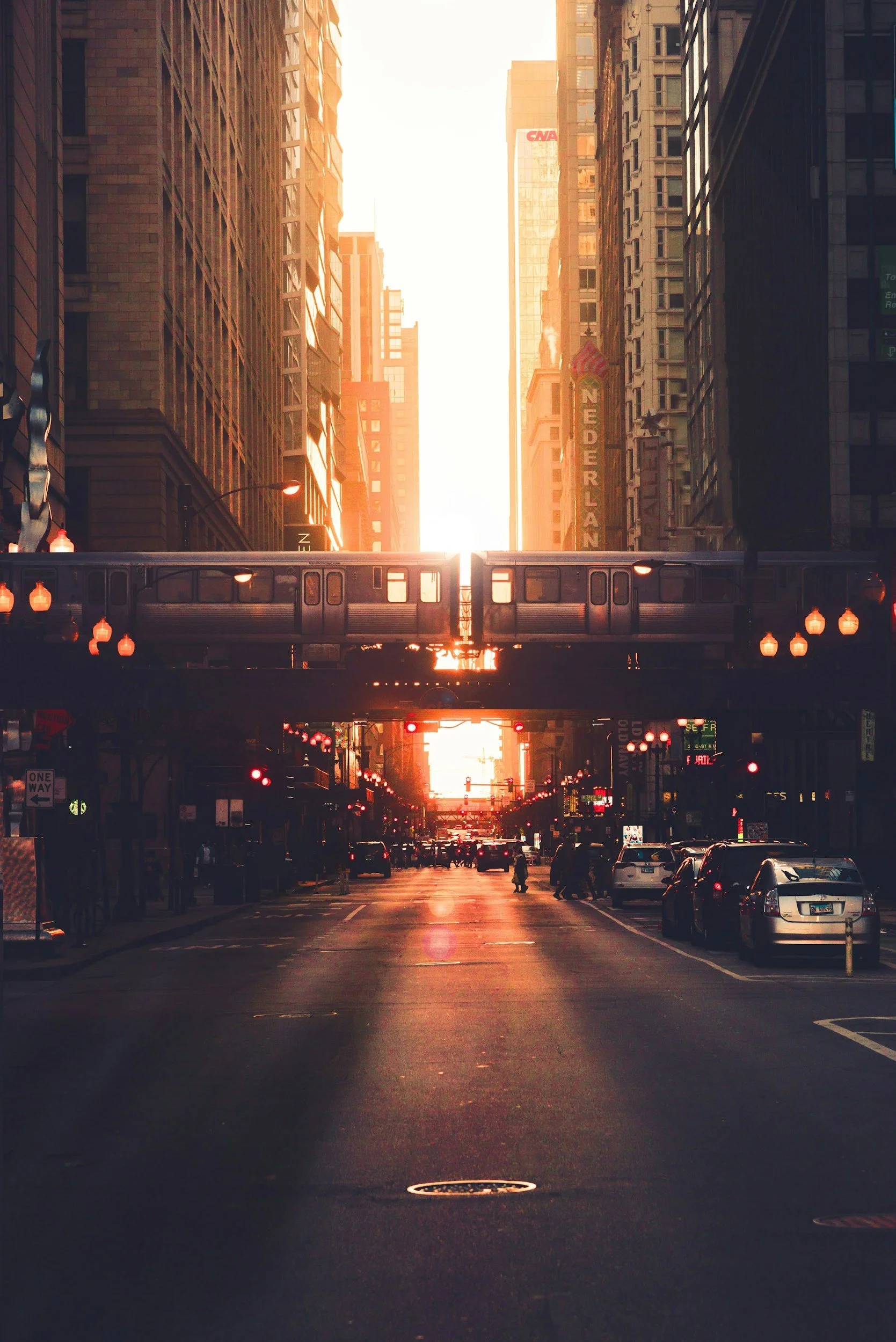 Street scene in a city with tall buildings on both sides, a train crossing above the street, cars parked along the street, and the sun setting in the background casting a warm glow over the scene.