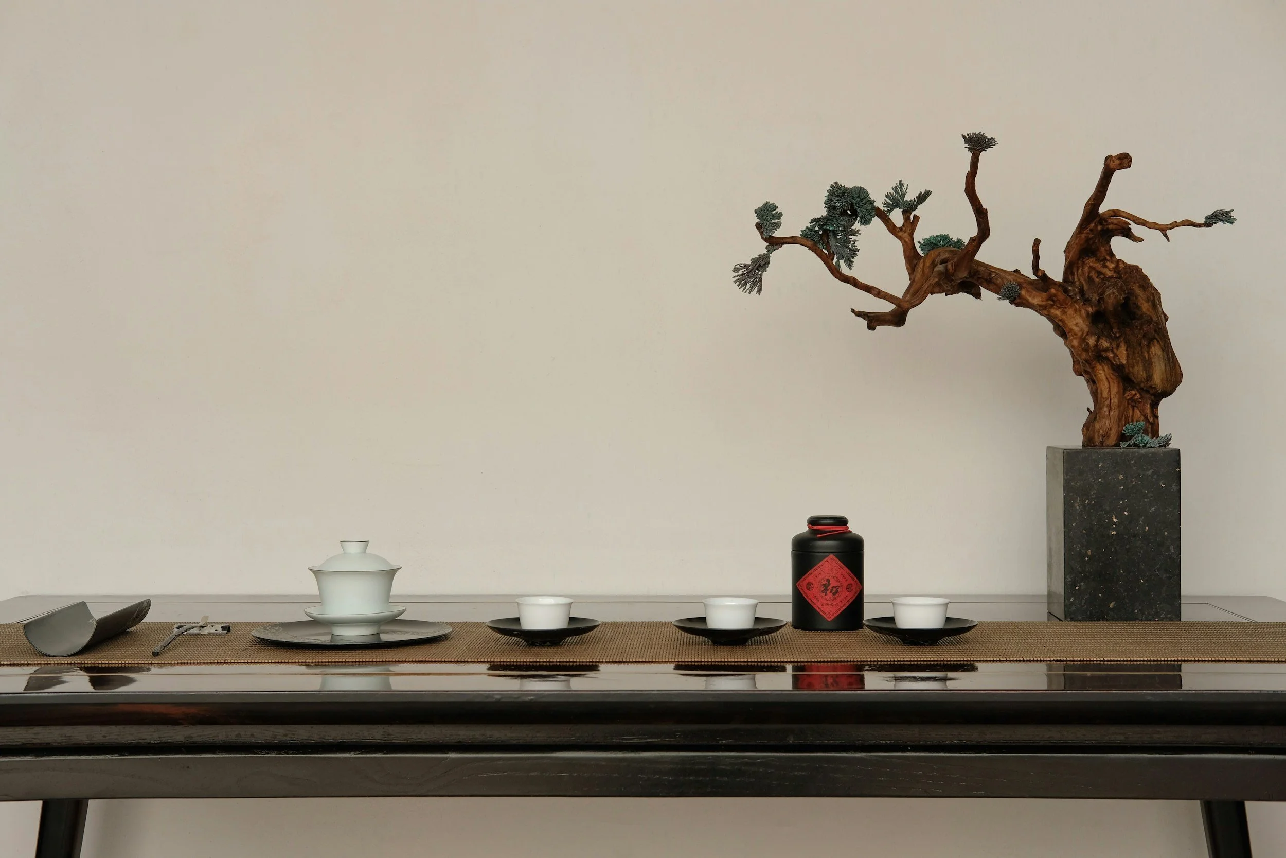 A minimalist setup on a table with a bonsai tree on a pedestal, traditional teacups, a black container with red Chinese characters, and a small white teapot, against a plain beige wall.