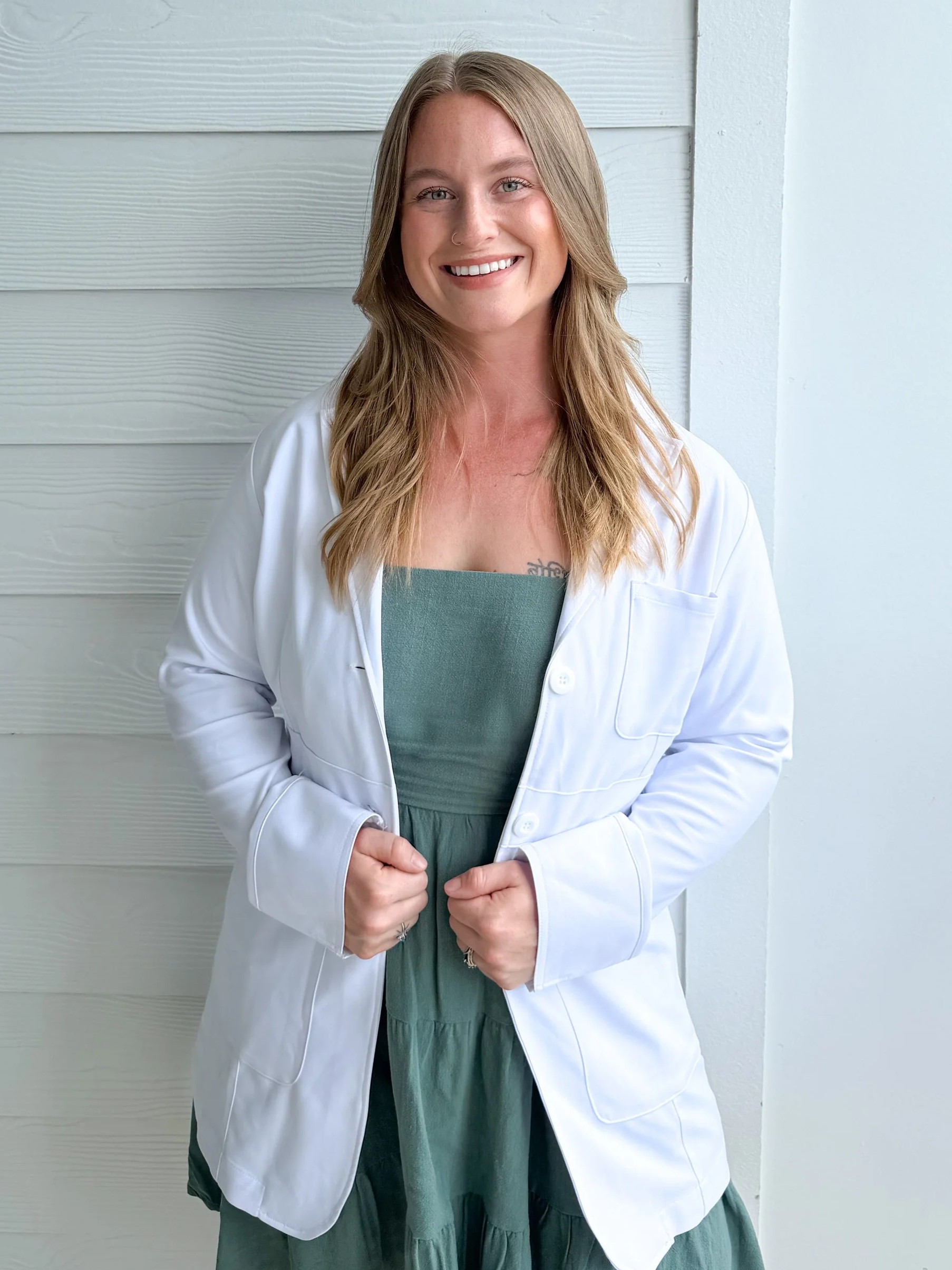 A smiling woman with blonde hair wearing a doctor's white coat and green dress standing against a white background.