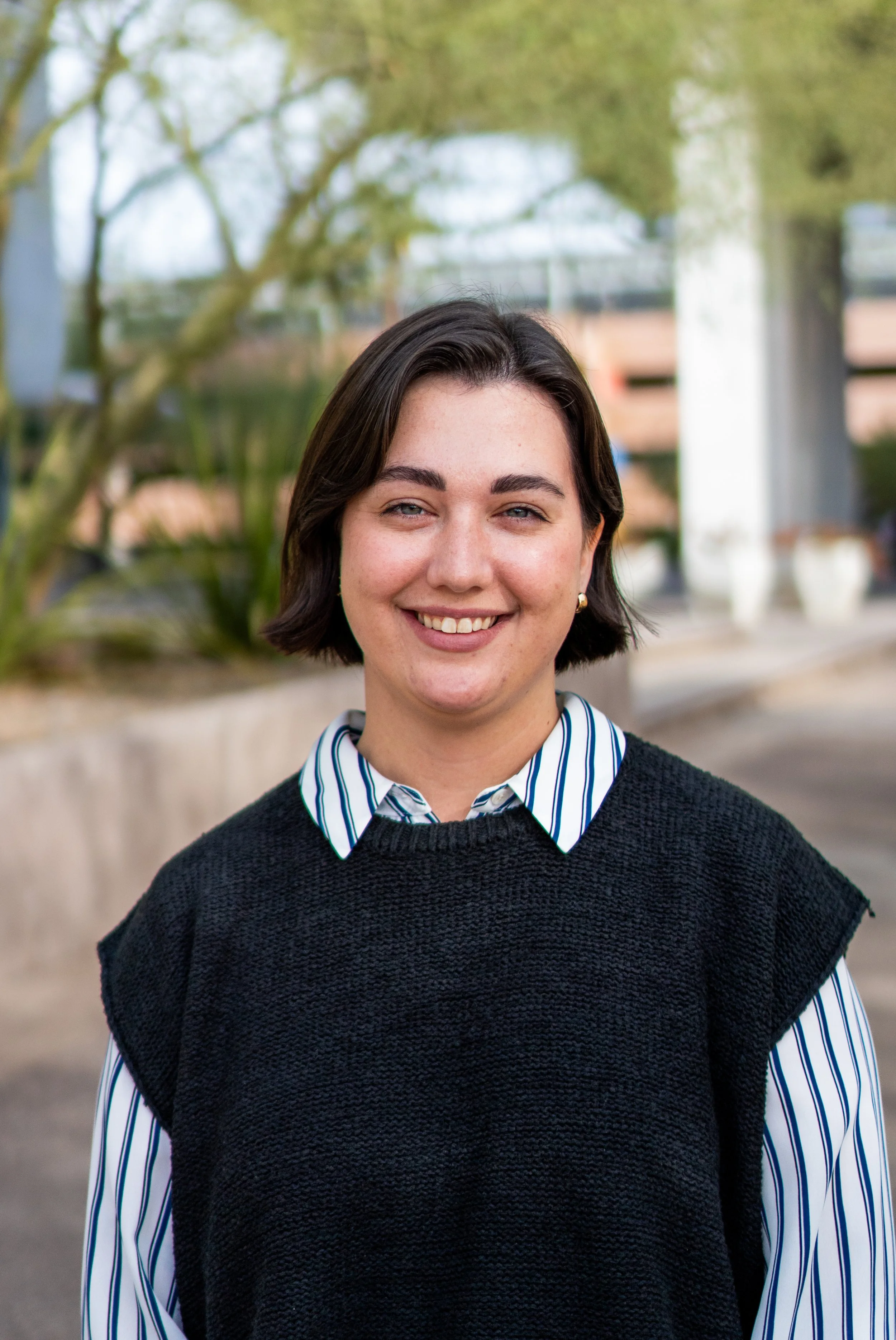 Close-up of a smiling woman with short dark hair, wearing a striped shirt under a black knitted vest, standing outdoors with trees and a building in the background.