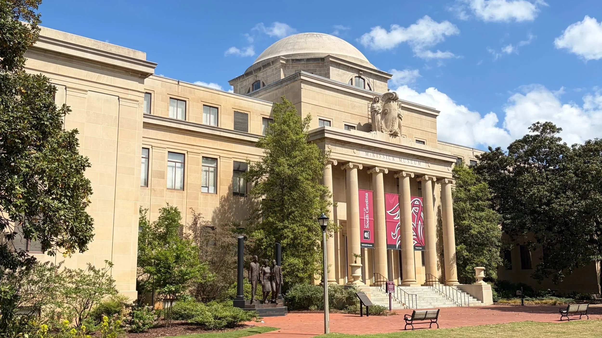The image shows the exterior of the C. McKissick Museum at the University of South Carolina, with classical architecture including columns and a dome. There are trees, benches, and statues in the foreground, and the sky is partly cloudy.