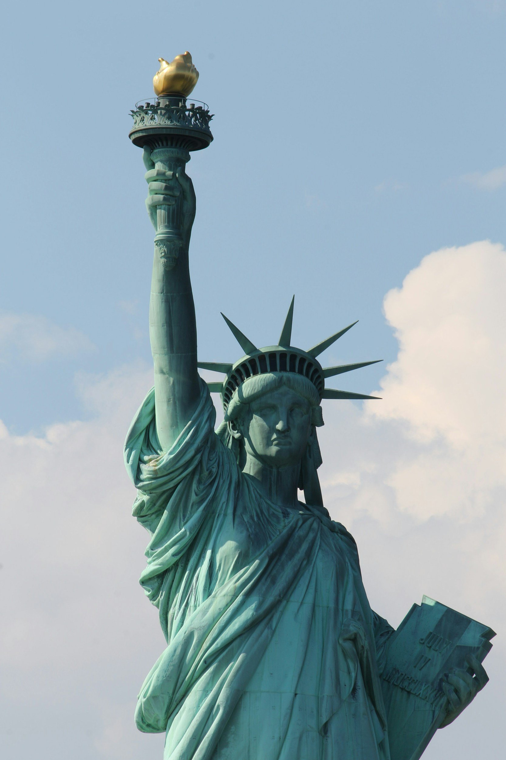 Close-up of the Statue of Liberty's face, torch, and part of the crown against a blue sky with a few clouds.