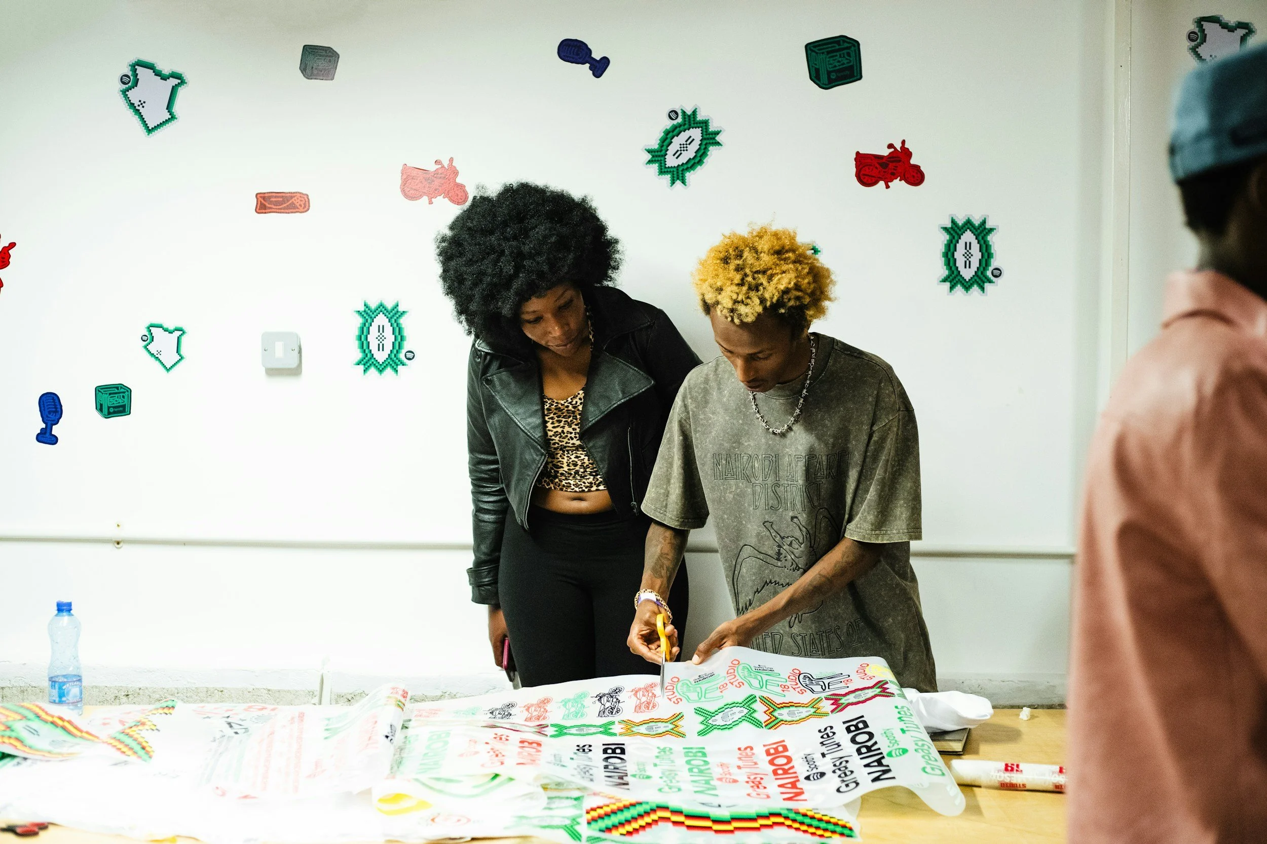 Two women stand at a table covered with colorful posters or fabrics with text and images. One woman has black curly hair and wears a black jacket, while the other has blonde curly hair and wears a casual gray T-shirt. The background wall displays various pixel art stickers, including motorcycles, cameras, and geometric shapes.