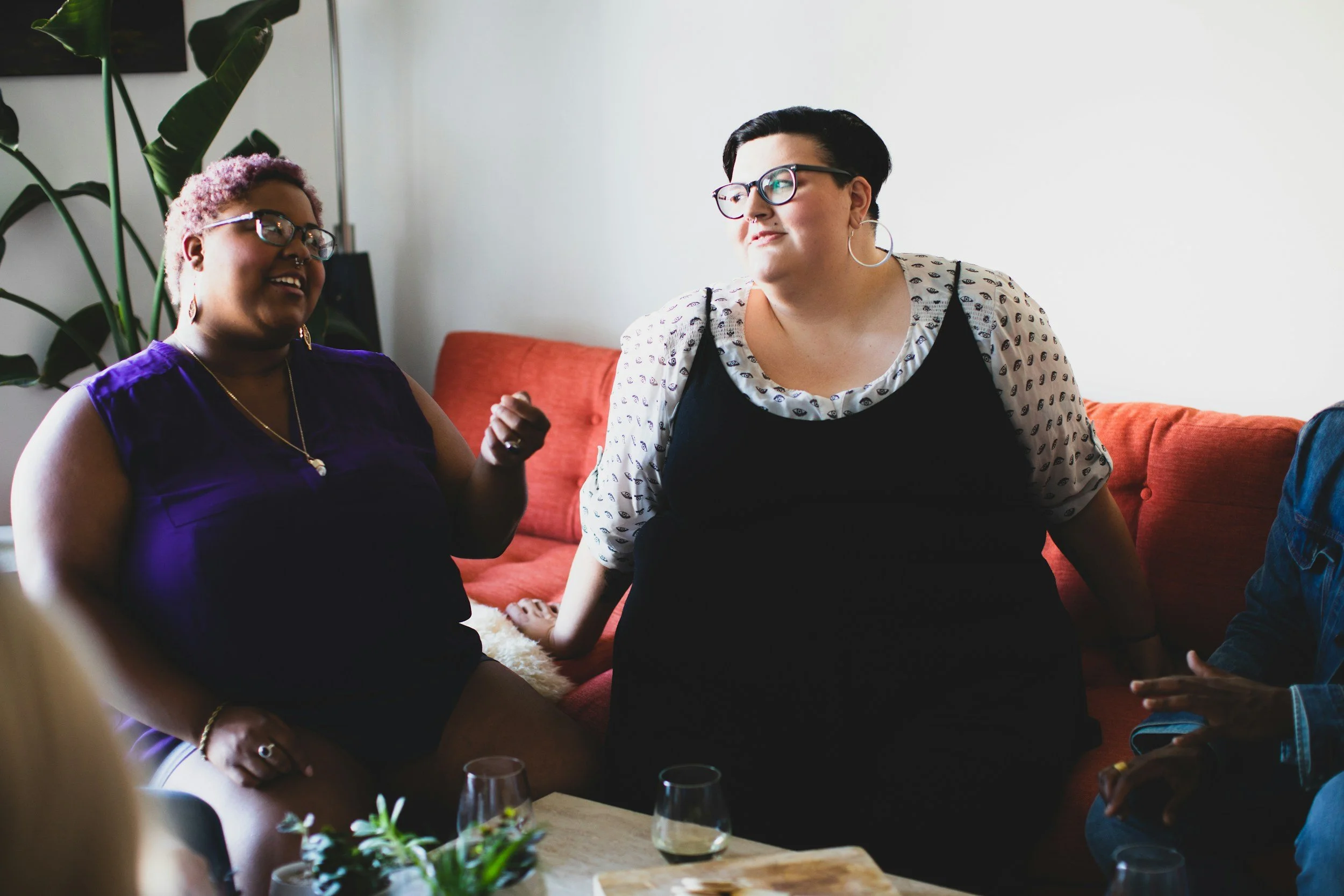 Two women sitting on a red couch, engaging in conversation inside a room with a white wall, with a large green plant behind them. One woman has short black hair, glasses, and earrings, wearing a white shirt with black dots and a black vest. The other woman has curly pink hair, glasses, and earrings, wearing a purple sleeveless top. There are drinks on a table in the foreground.