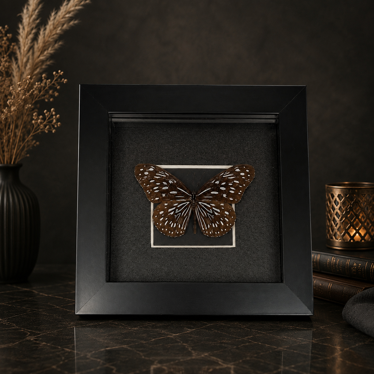 Framed butterfly display with brown and white wings, placed on a black background with decorative items like a vase with dried plants and a candle holder on a dark marble surface.