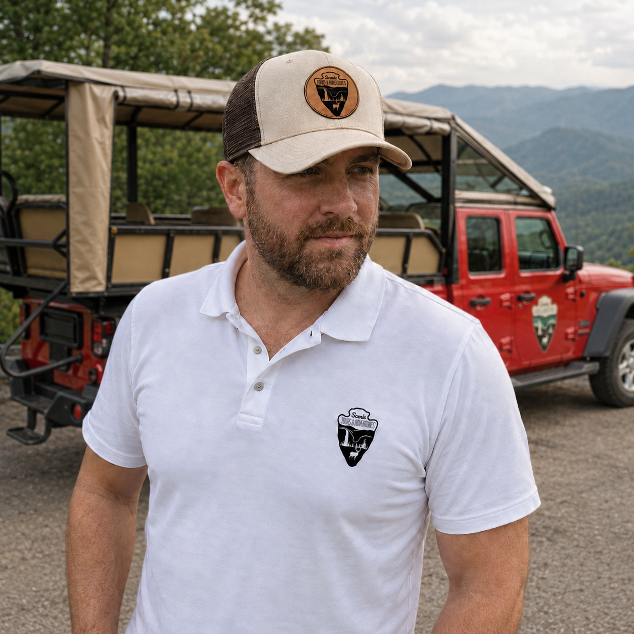 A man wearing a beige and brown cap and a white polo shirt with a logo, standing outdoors with a red safari vehicle and mountainous landscape in the background.