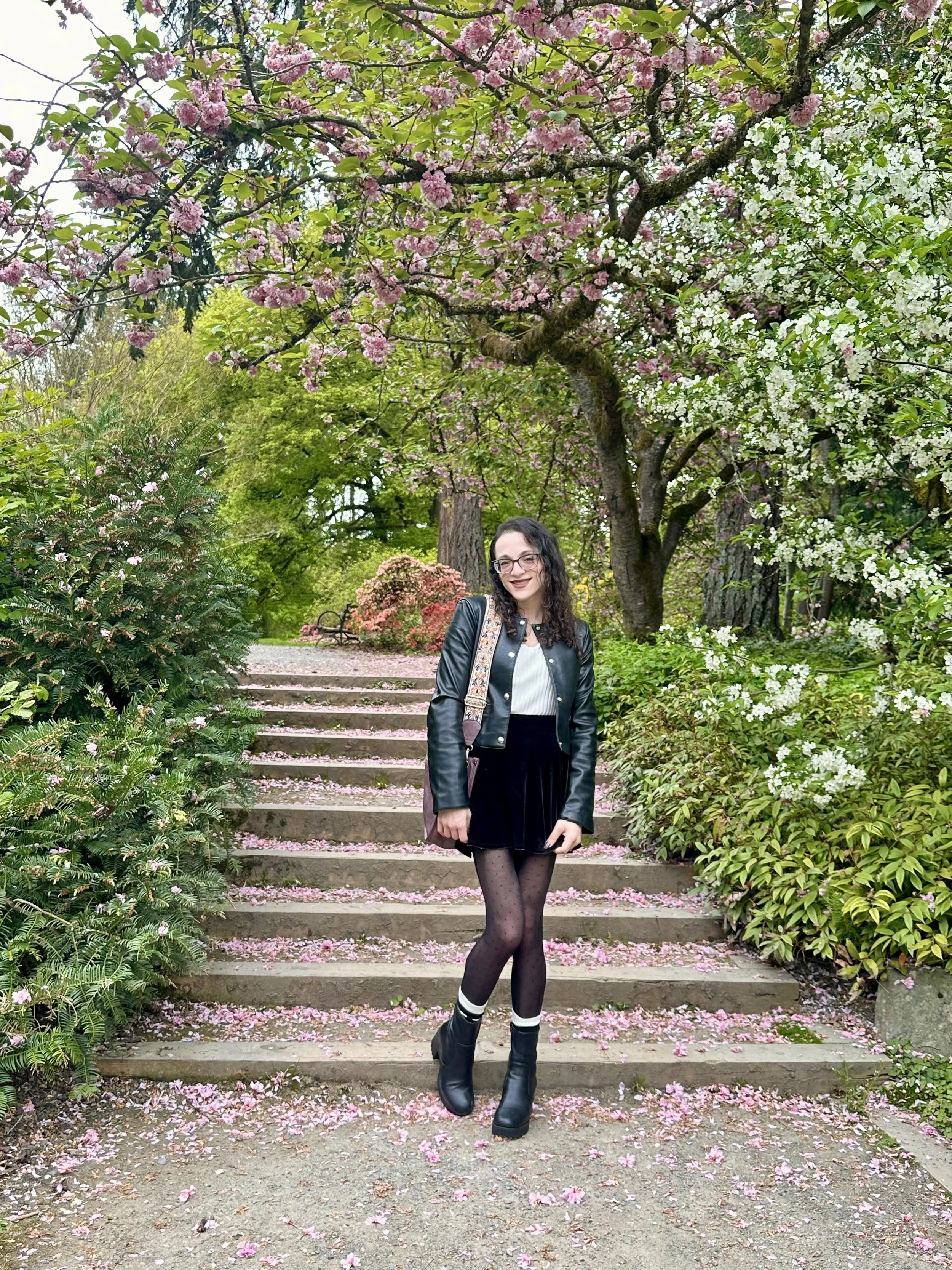 A woman (Leslie) with glasses and long curly hair standing on outdoor steps covered in pink flower petals, surrounded by blooming trees and green foliage.