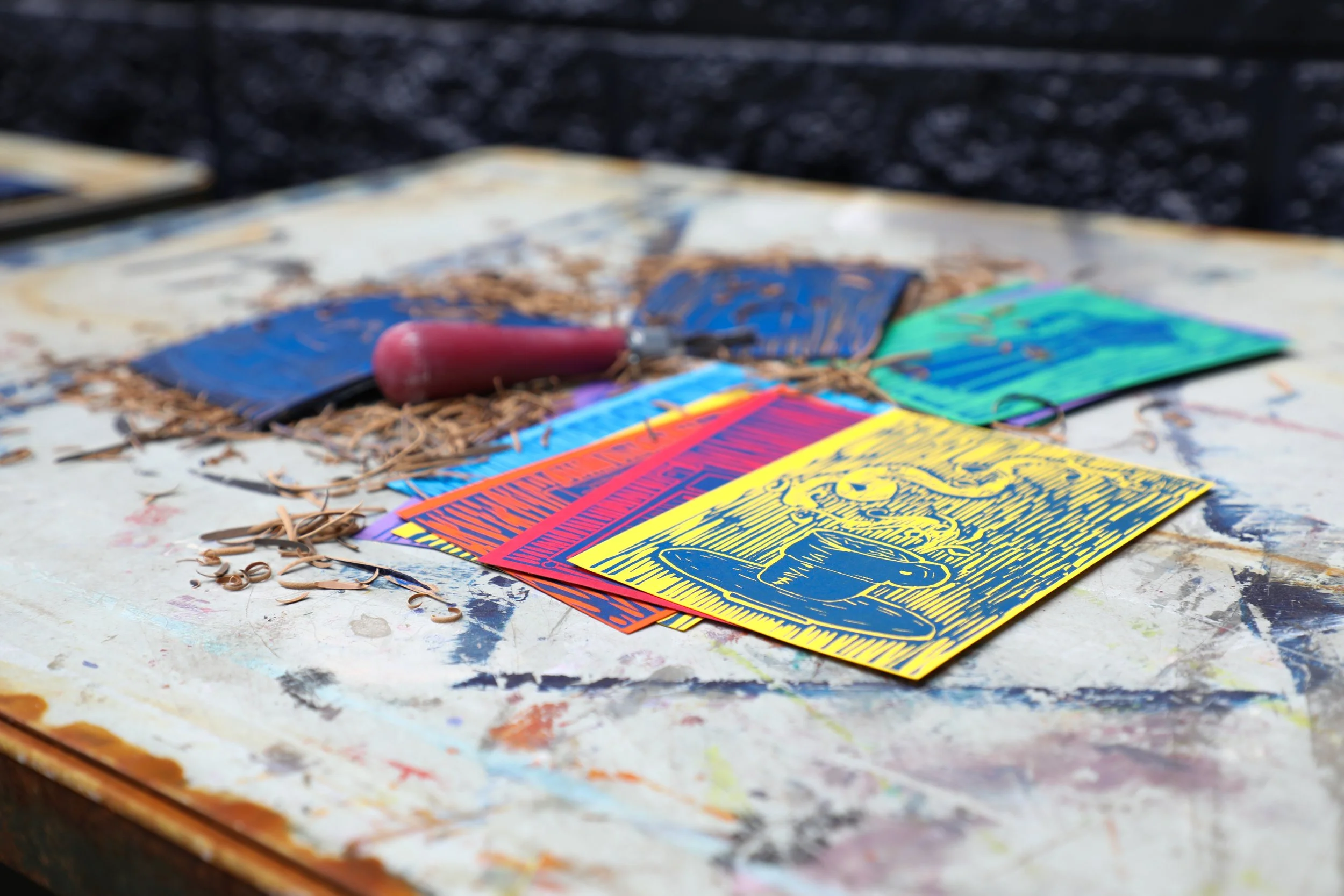 Colorful flyers or posters with artistic designs and text lying on a paint-splattered table. Art tools and scattered shredded pieces of linolium are also visible.