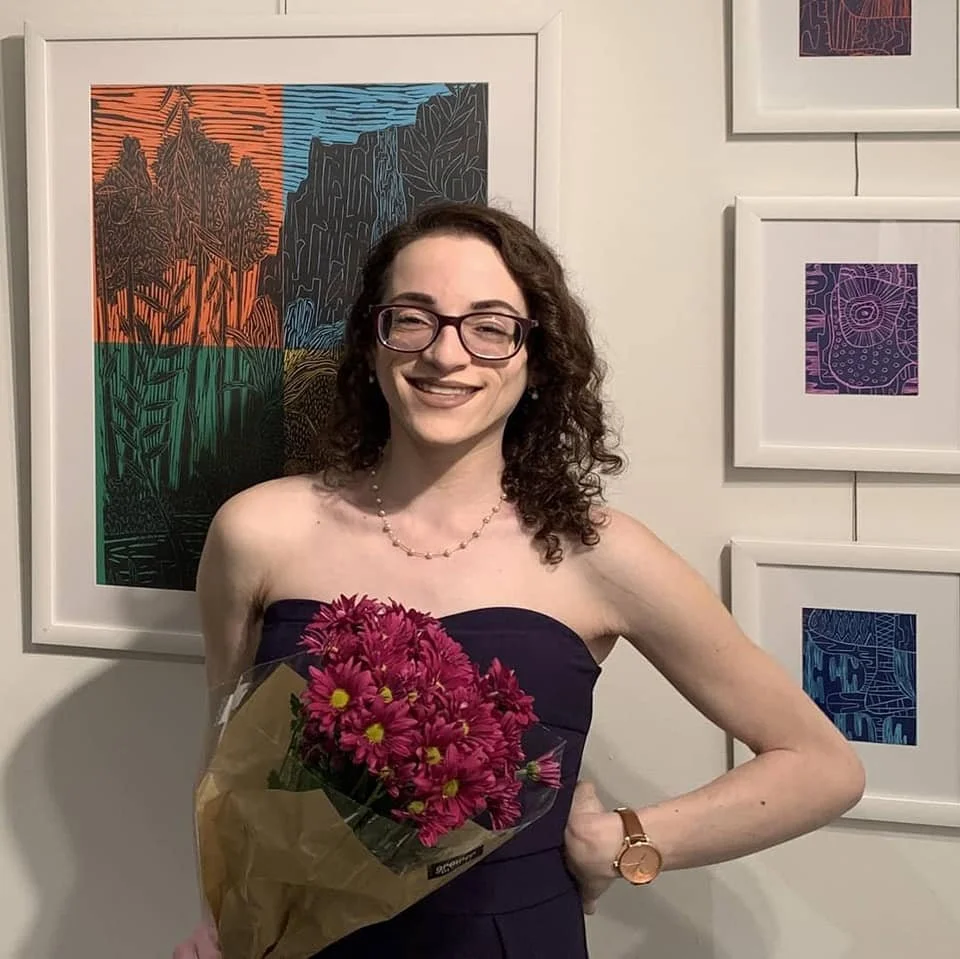 A woman (Leslie) with curly brown hair, glasses, and a strapless dark dress is holding a bouquet of pink flowers and smiling. She stands in front of a wall with framed abstract and colorful artwork.