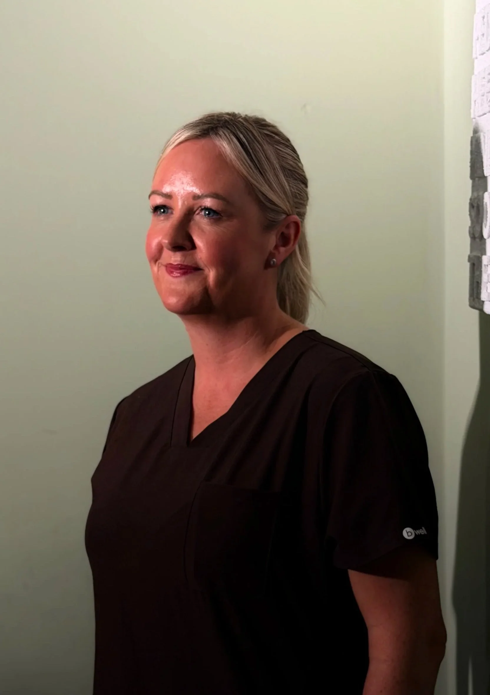 A woman with blonde hair tied back wearing a black medical uniform, standing against a light green wall, smiling slightly.