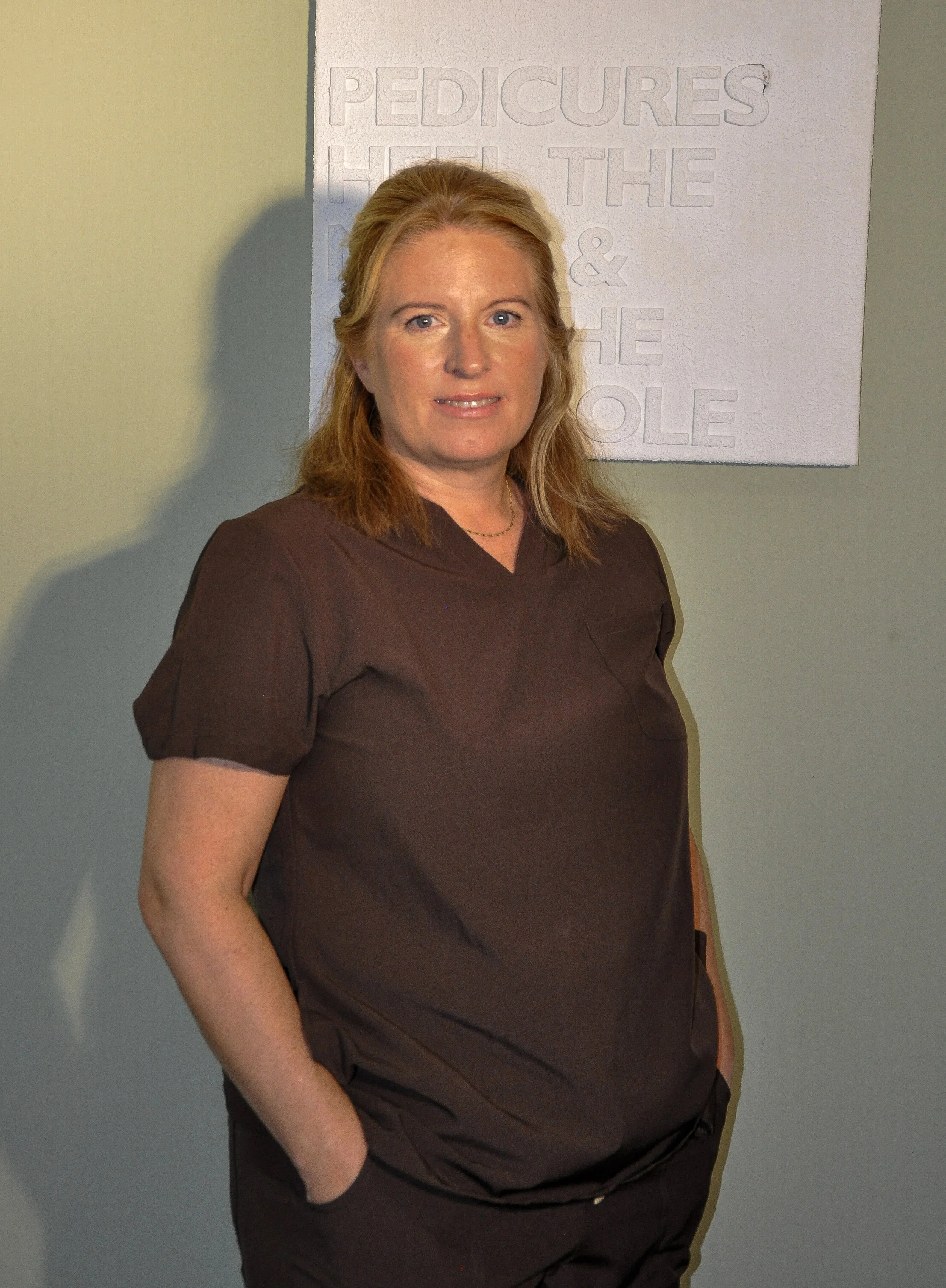 A woman with shoulder-length blonde hair, wearing a dark brown top, stands in front of a white textured wall with the embossed words 'PEDICURES HEAL THE & THE OLE'. She has a slight smile and is looking at the camera.
