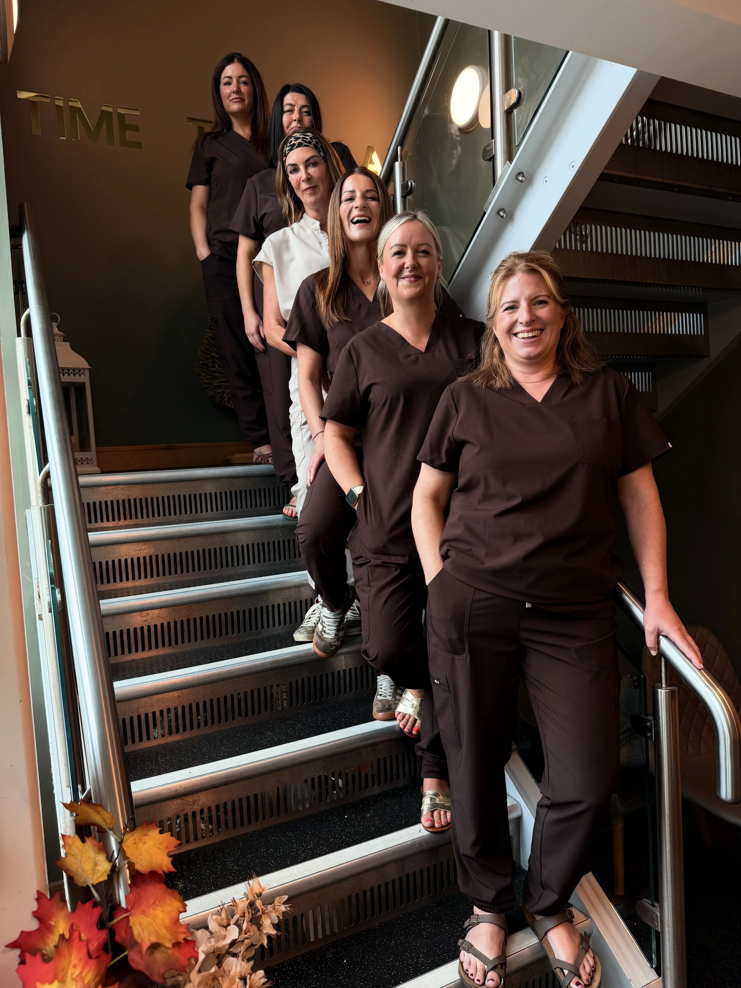 Six women in brown and white scrubs standing on a staircase, posing for a photo, with autumn leaves decoration at the bottom.
