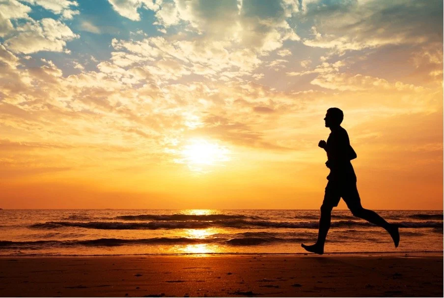Silhouette of a person jogging on the beach during a sunset with orange and yellow sky and calm ocean waves.