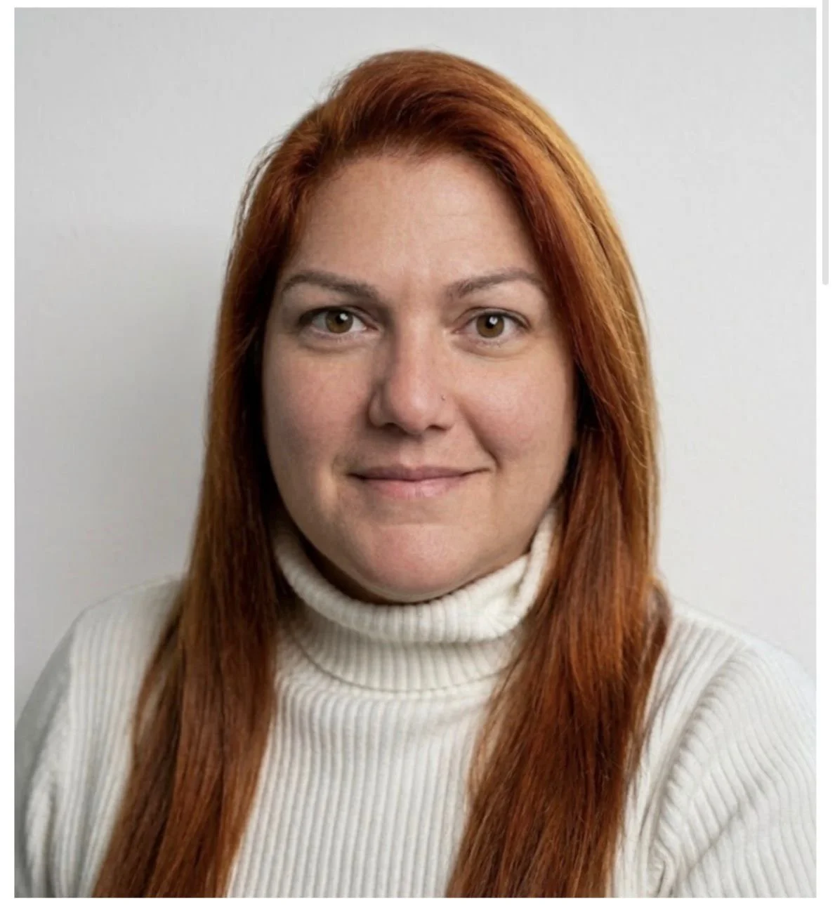 A woman with long red hair, fair skin, and hazel eyes, wearing a white turtleneck sweater, smiling slightly against a plain white background.