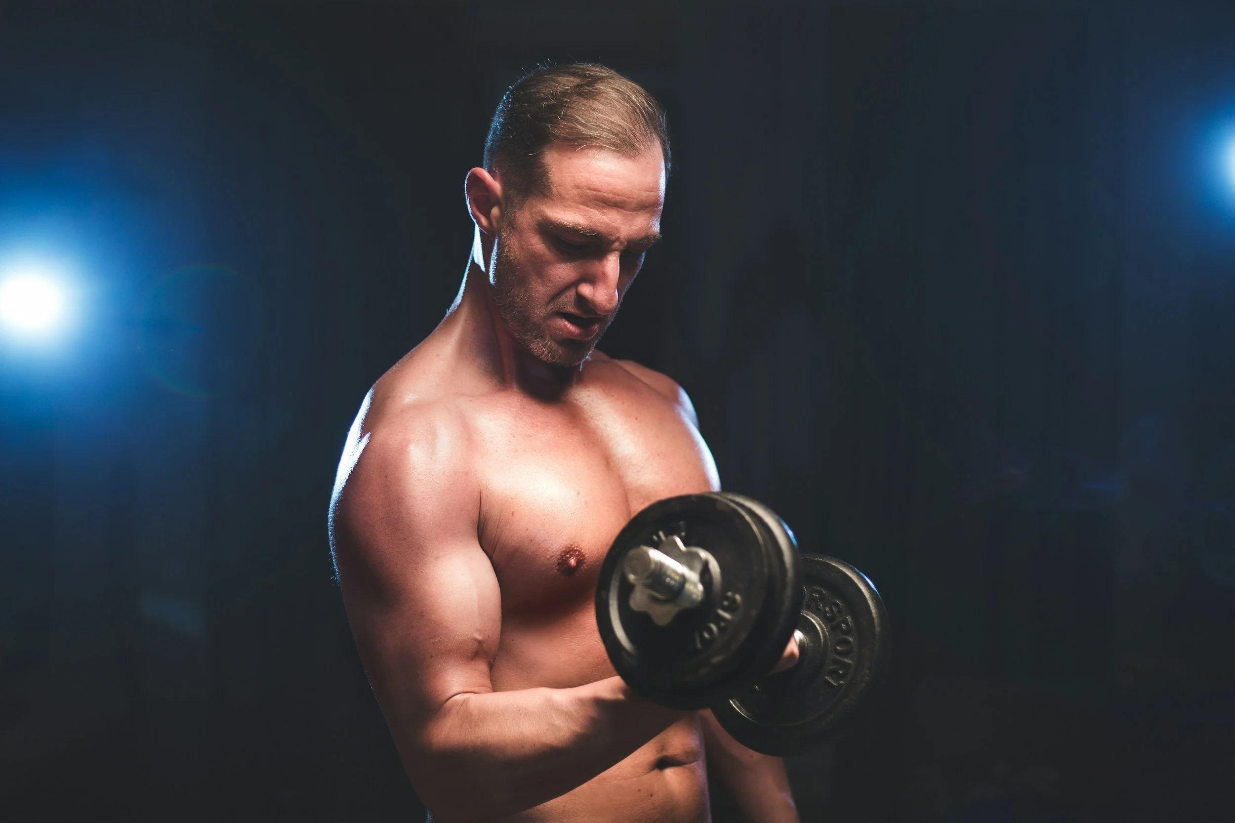 A shirtless man that needs hormone replacement therapy with short hair and a beard working out with dumbbells in a dark gym setting, lit by two bright lights on either side.