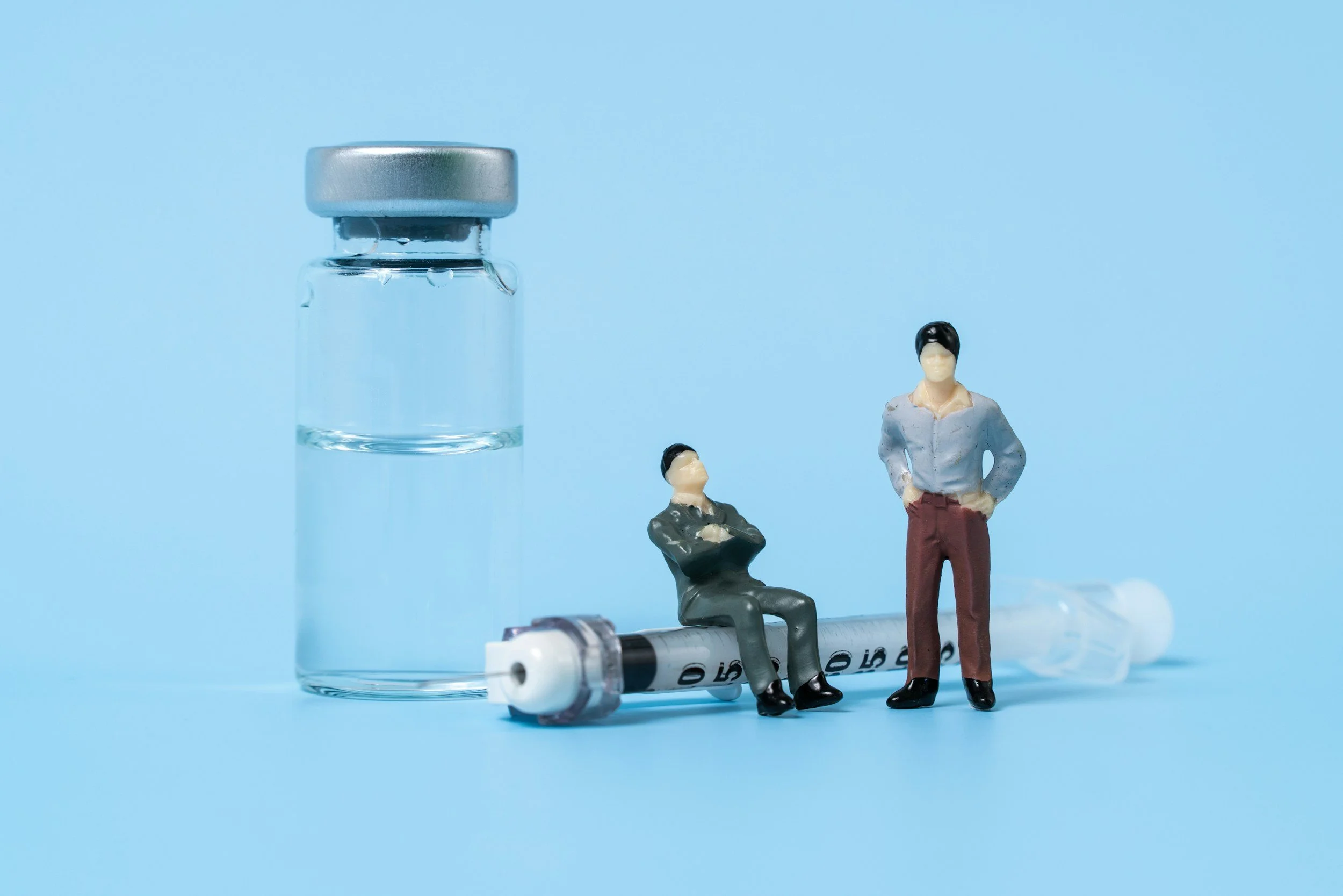 A clear Peptide glass vial with a metal lid, a syringe with a needle, and three miniature figurines of people against a light blue background.