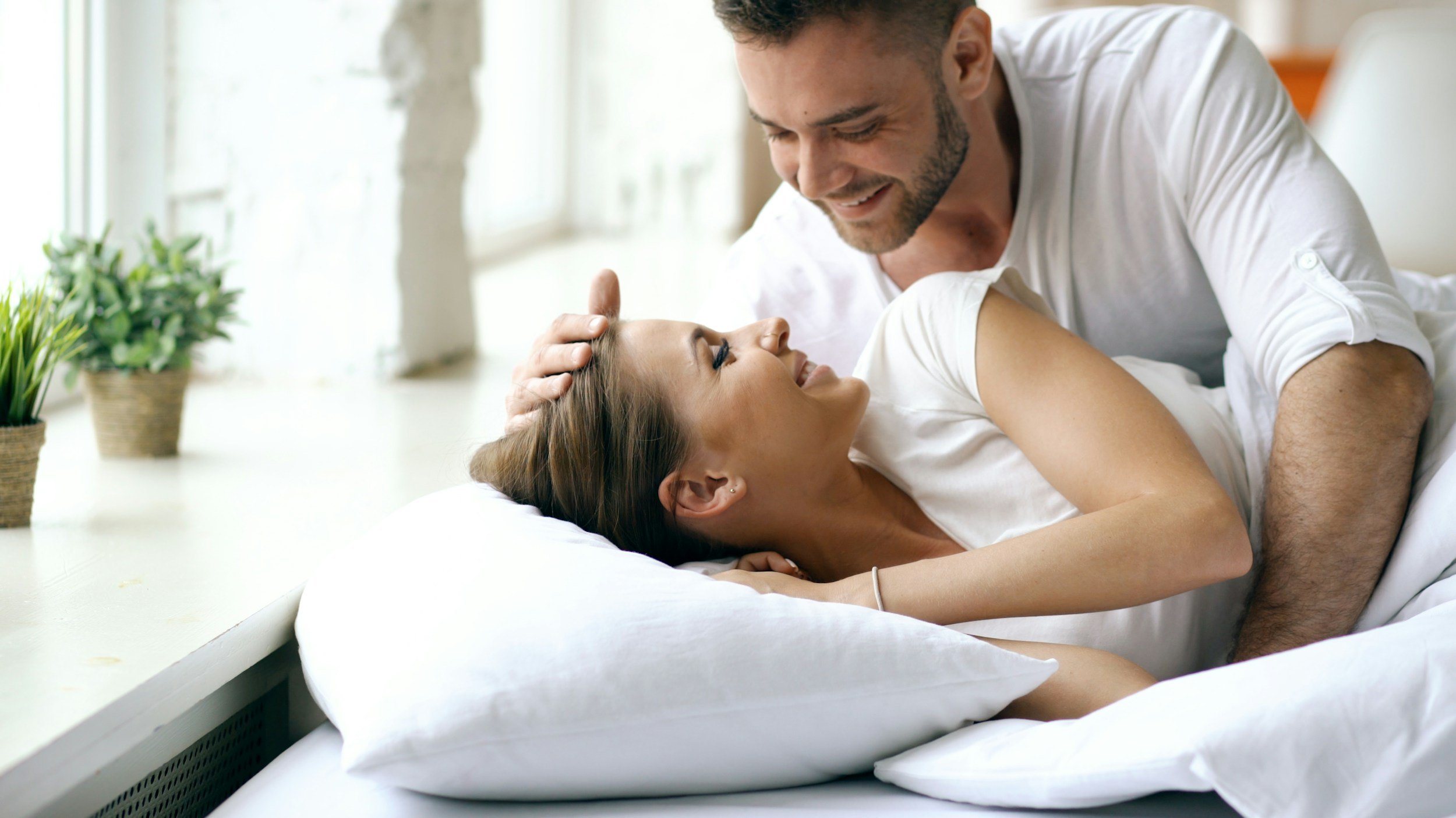 A happy couple sharing a moment in bed, with the man gently touching the woman's head and both smiling warmly at each other in a bright, cozy room.