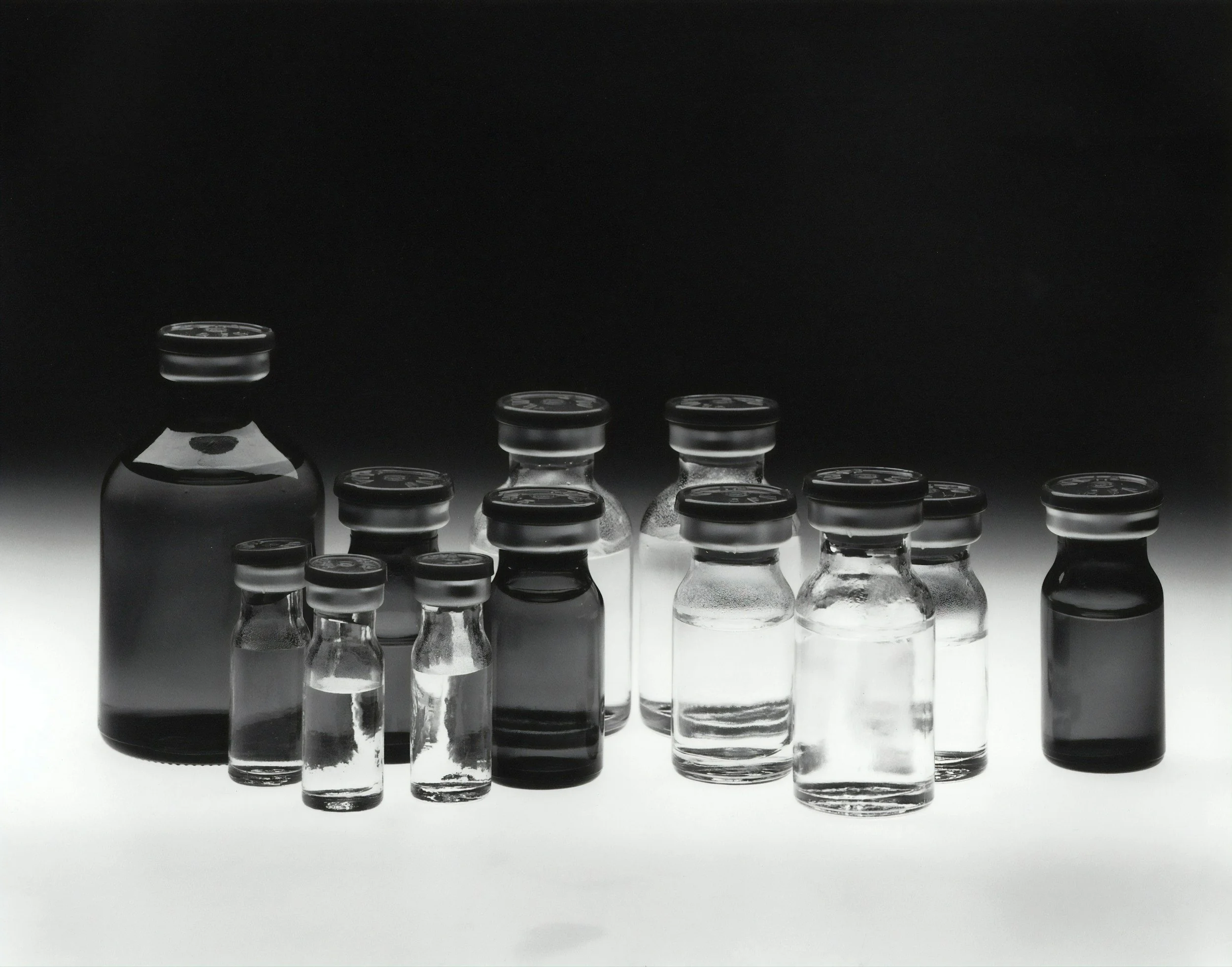 A row of glass vials with black caps, arranged on a reflective surface, with a black background.
