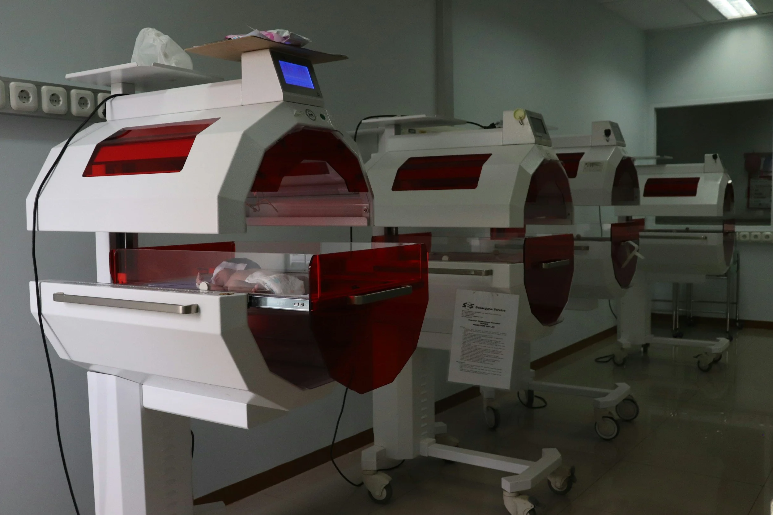 Medical incubators with transparent red covers on medical carts in a hospital.