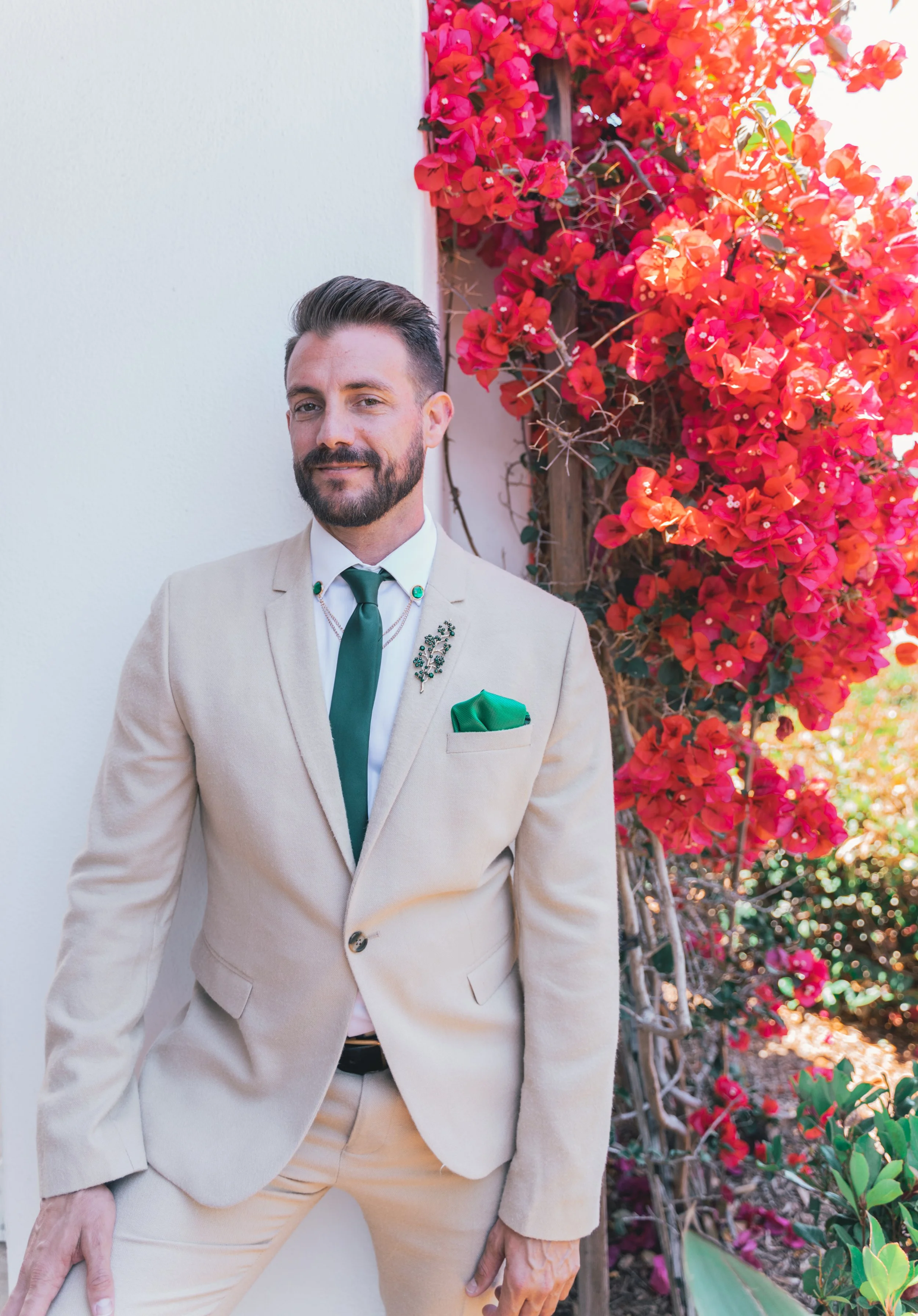 A functional medicine Doctor in Miami man with a beard and styled hair wearing a beige suit, white shirt, and dark green tie, standing outdoors next to a wall with vibrant pink and red bougainvillea flowers in the background.
