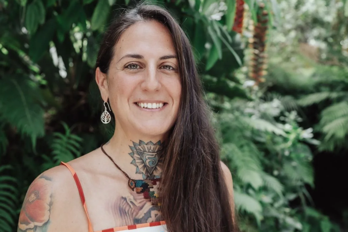 A smiling woman with long dark hair and tattoos on her arm and neck, wearing earrings and a necklace, standing outdoors among green plants.