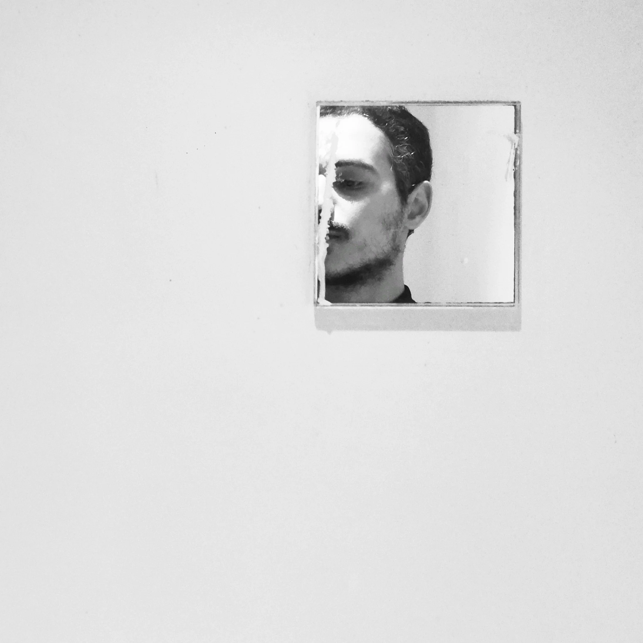 A black and white photo of a person with curly hair seen through a mirror in a plain room.