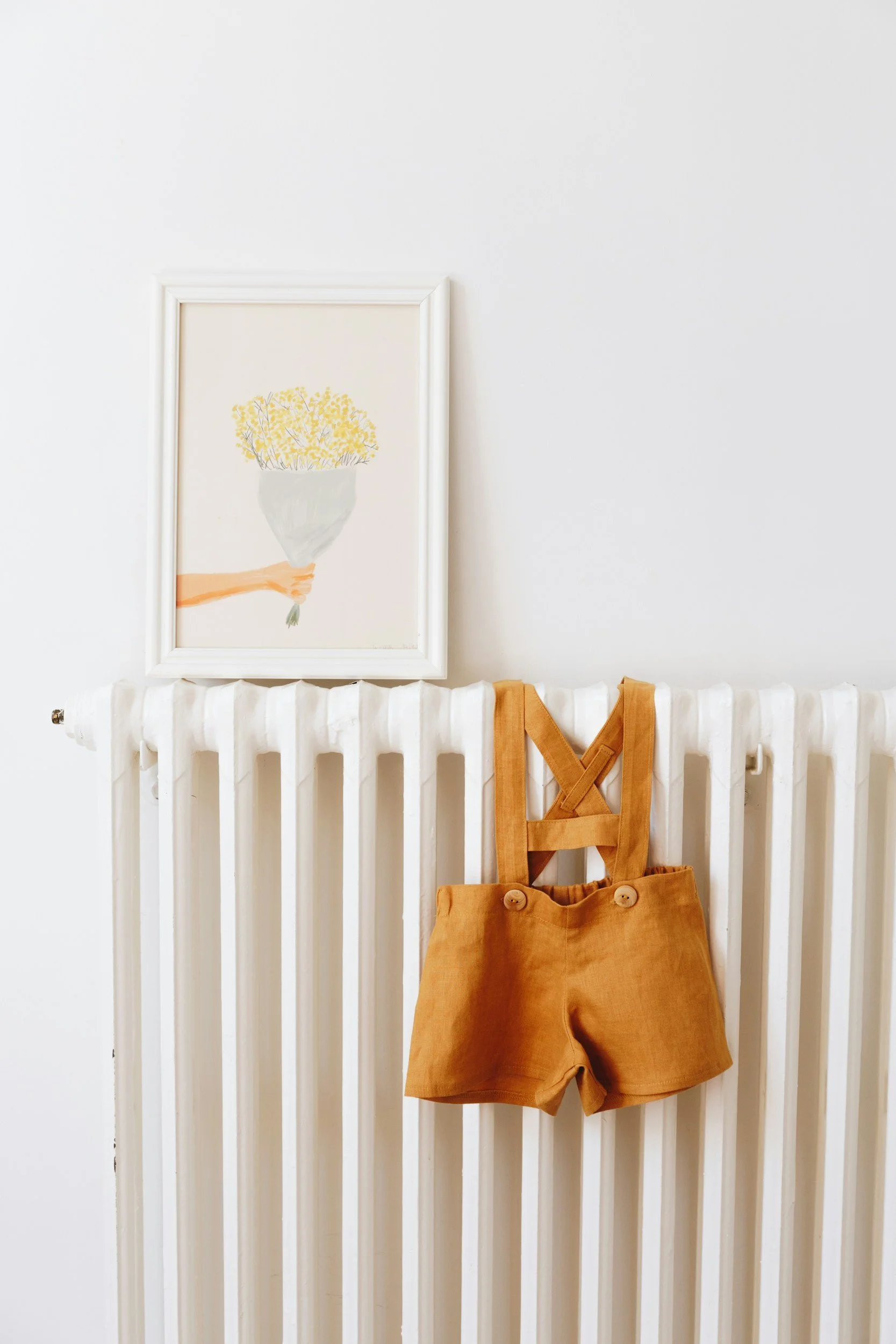 A white radiator mounted on a white wall with a framed watercolor painting of a yellow floral arrangement resting on top. A small orange-brown apron hangs from the radiator.