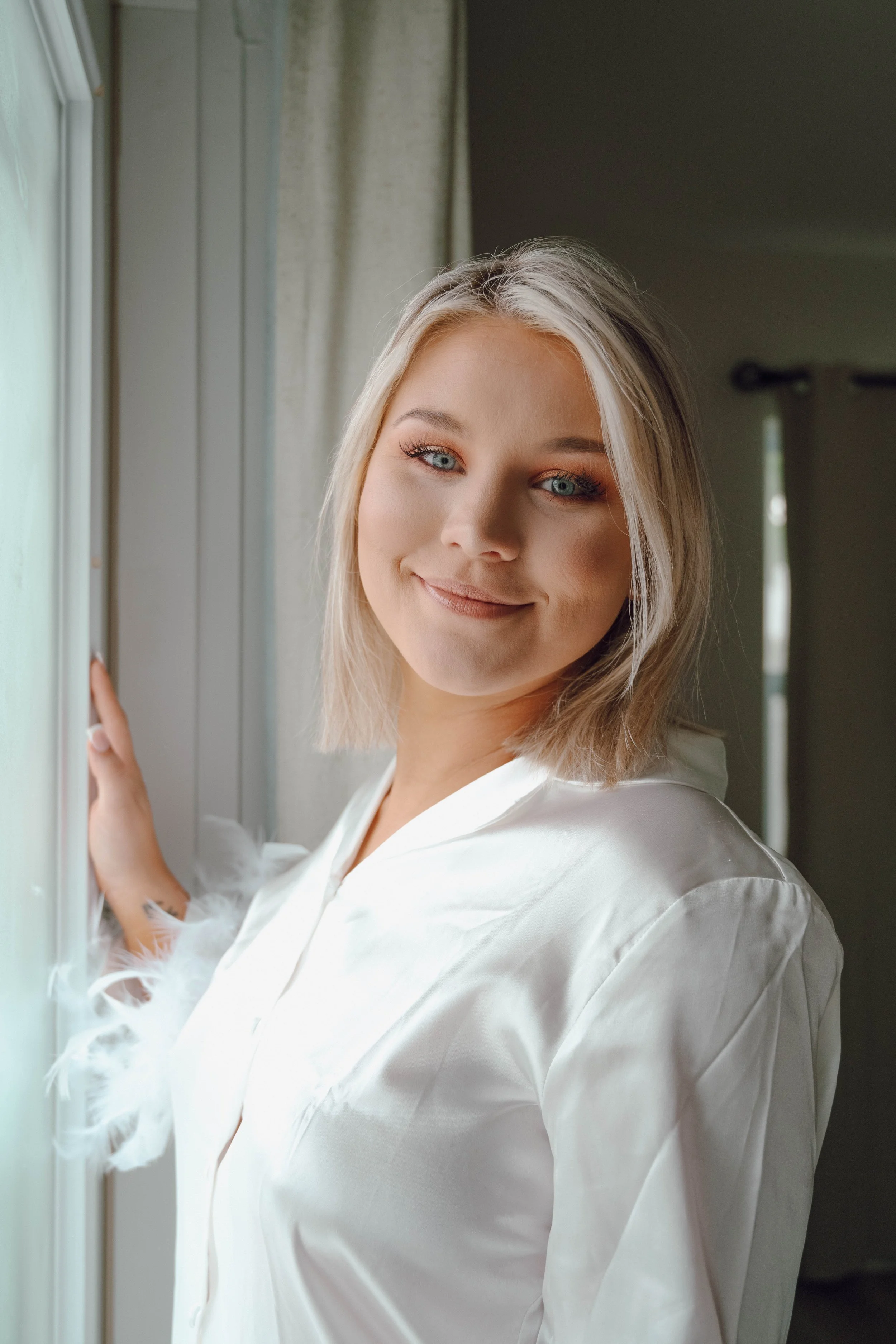 A young woman with long blonde hair, light skin, and blue eyes, smiling gently while looking at the camera. She is wearing a white shirt with feathered details on the sleeves and stands indoors near a window with beige curtains.