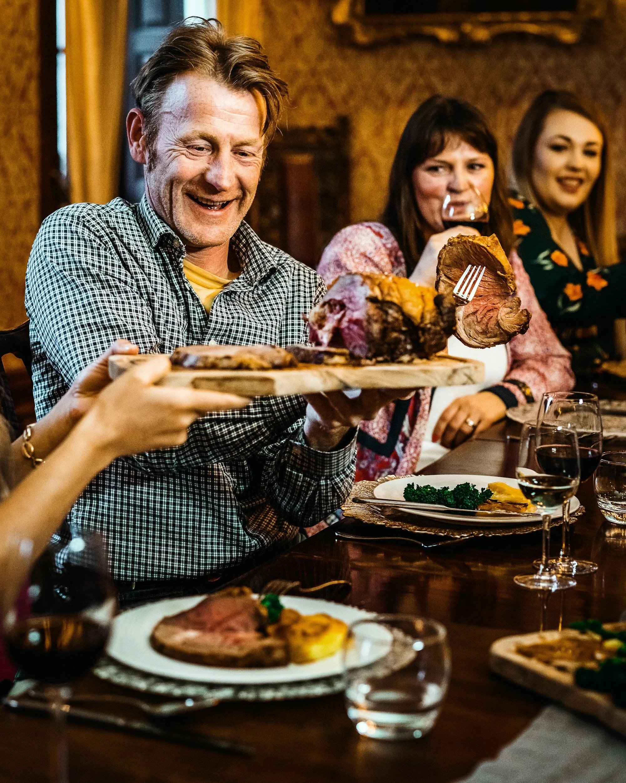 People enjoying a meal at a restaurant, with a man smiling and holding a plate of roast beef, other diners in the background, and a table with various dishes and glasses of wine.