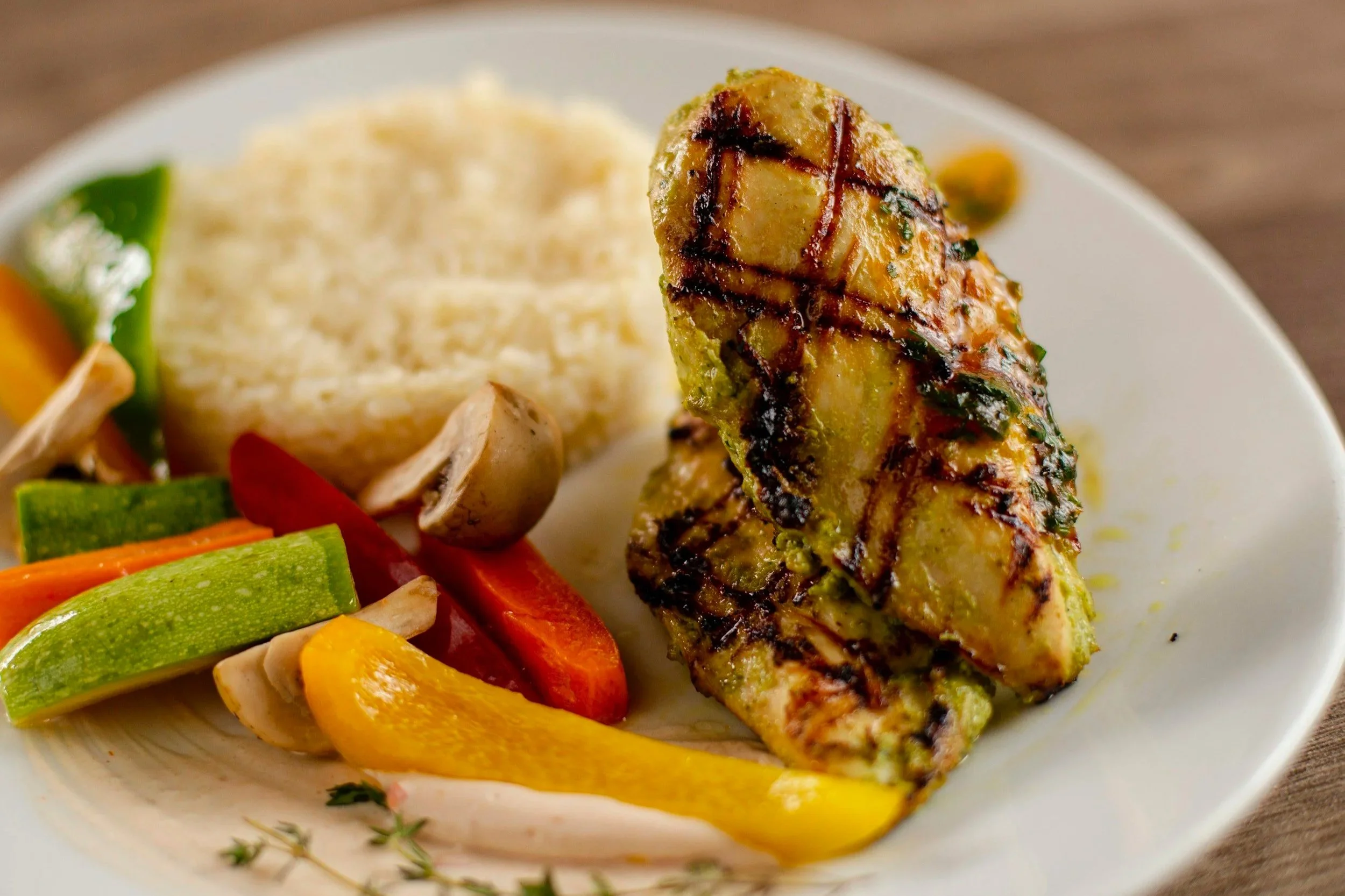 Grilled chicken leg quarter with grill marks, served with roasted vegetables including red and yellow bell peppers, carrots, zucchini, and mushrooms, and a scoop of mashed potatoes.