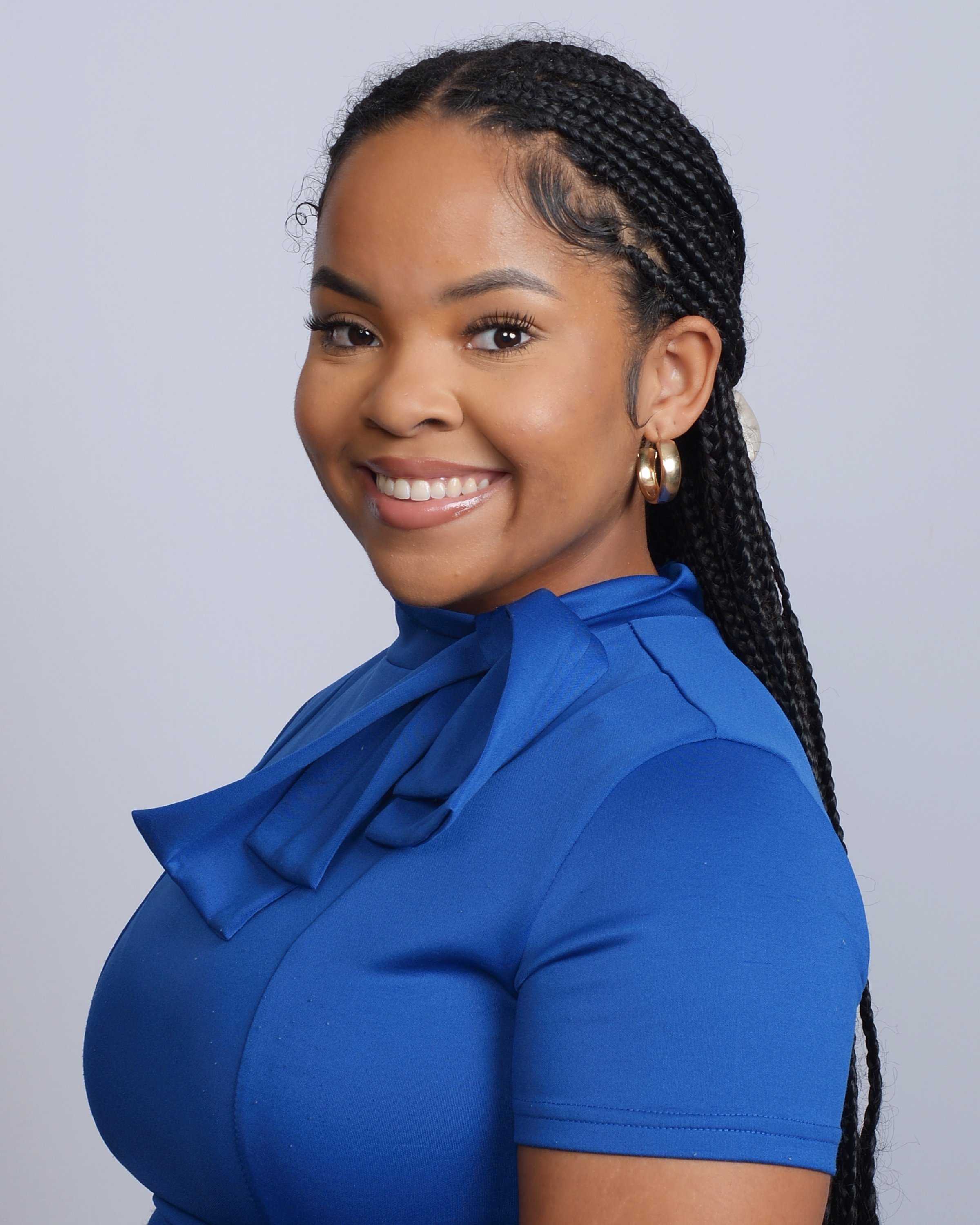 Close-up of a smiling woman with braided hair, wearing a blue dress with a ruffled collar and large gold hoop earrings, against a plain light gray background.