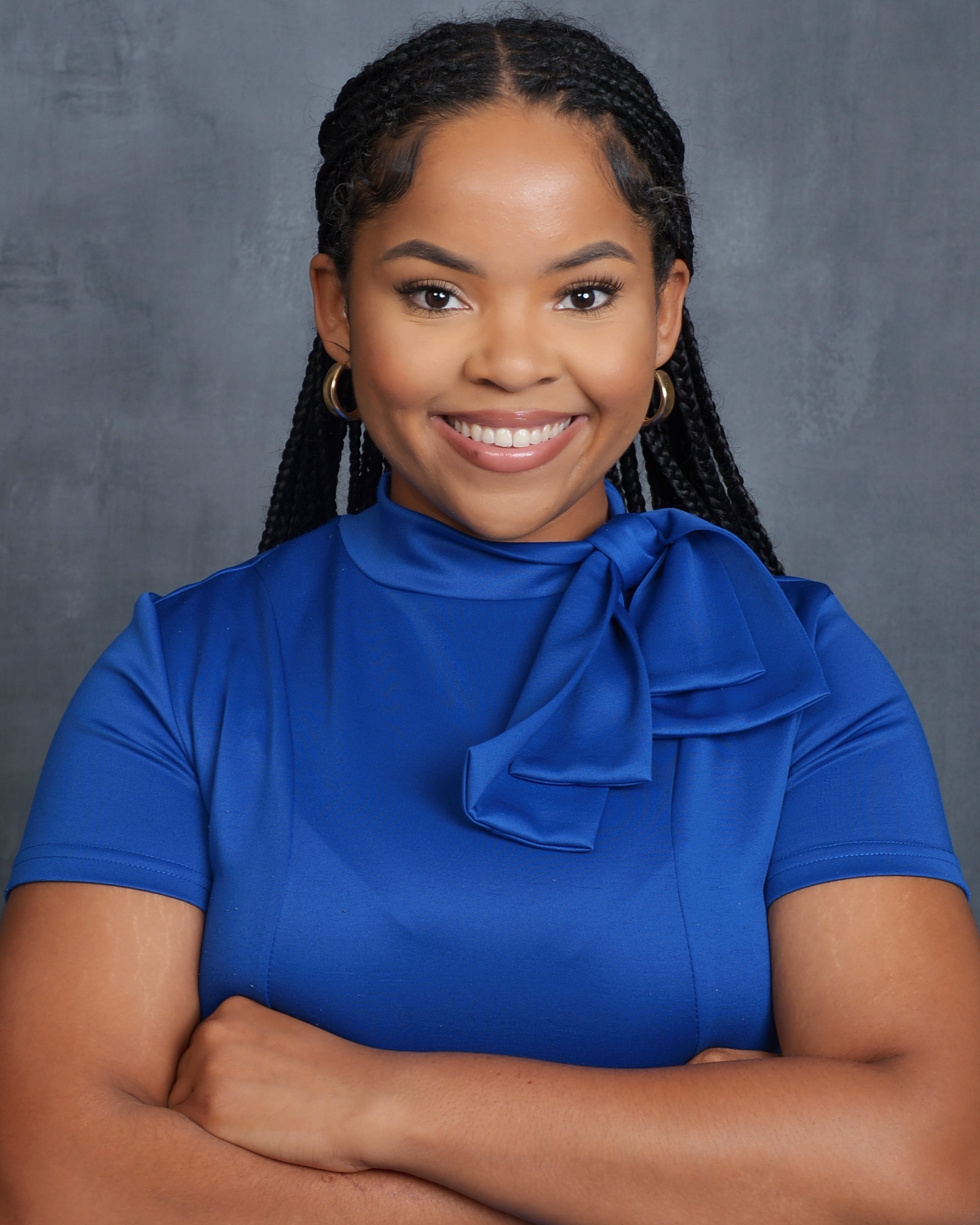 Close-up of a woman smiling with arms crossed, wearing a bright blue top and gold hoop earrings, standing against a gray background.