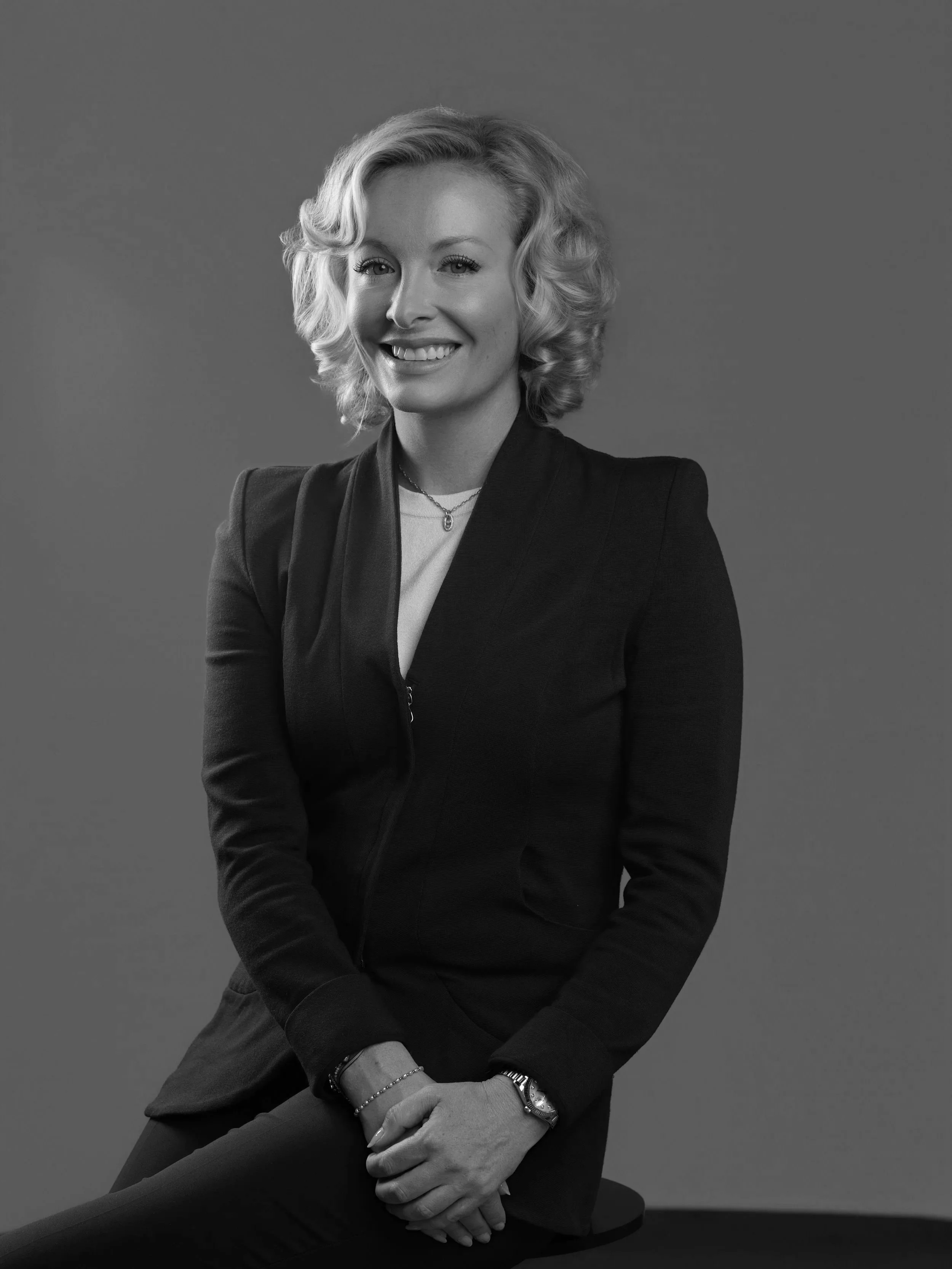 A black and white portrait of Natasha French, smiling, dressed in professional attire, wearing a necklace, watch, and bracelets, sitting against a plain background.
