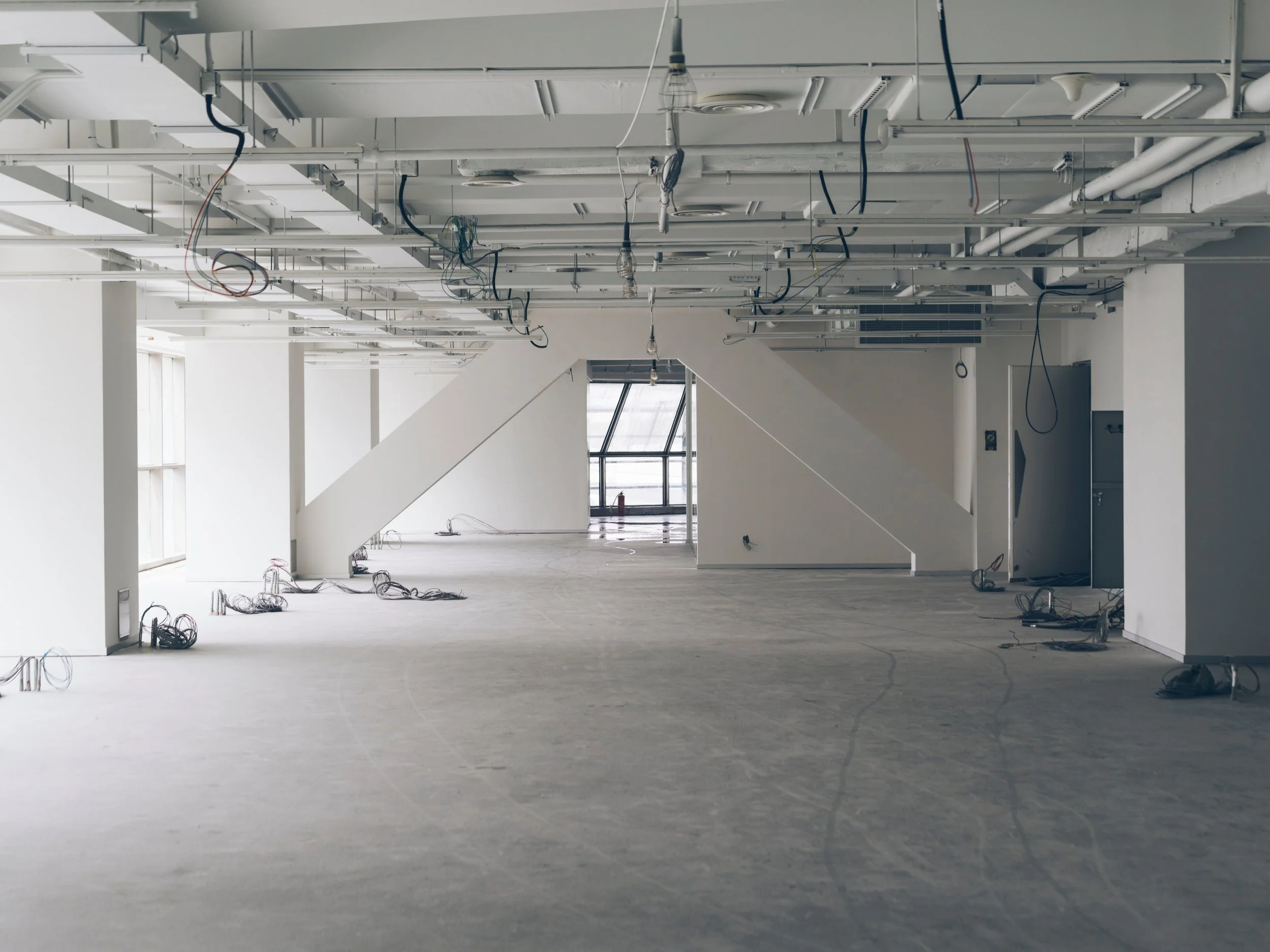 An unfinished interior space with exposed ceiling pipes, wiring, and hanging light bulbs, with large windows and white walls.