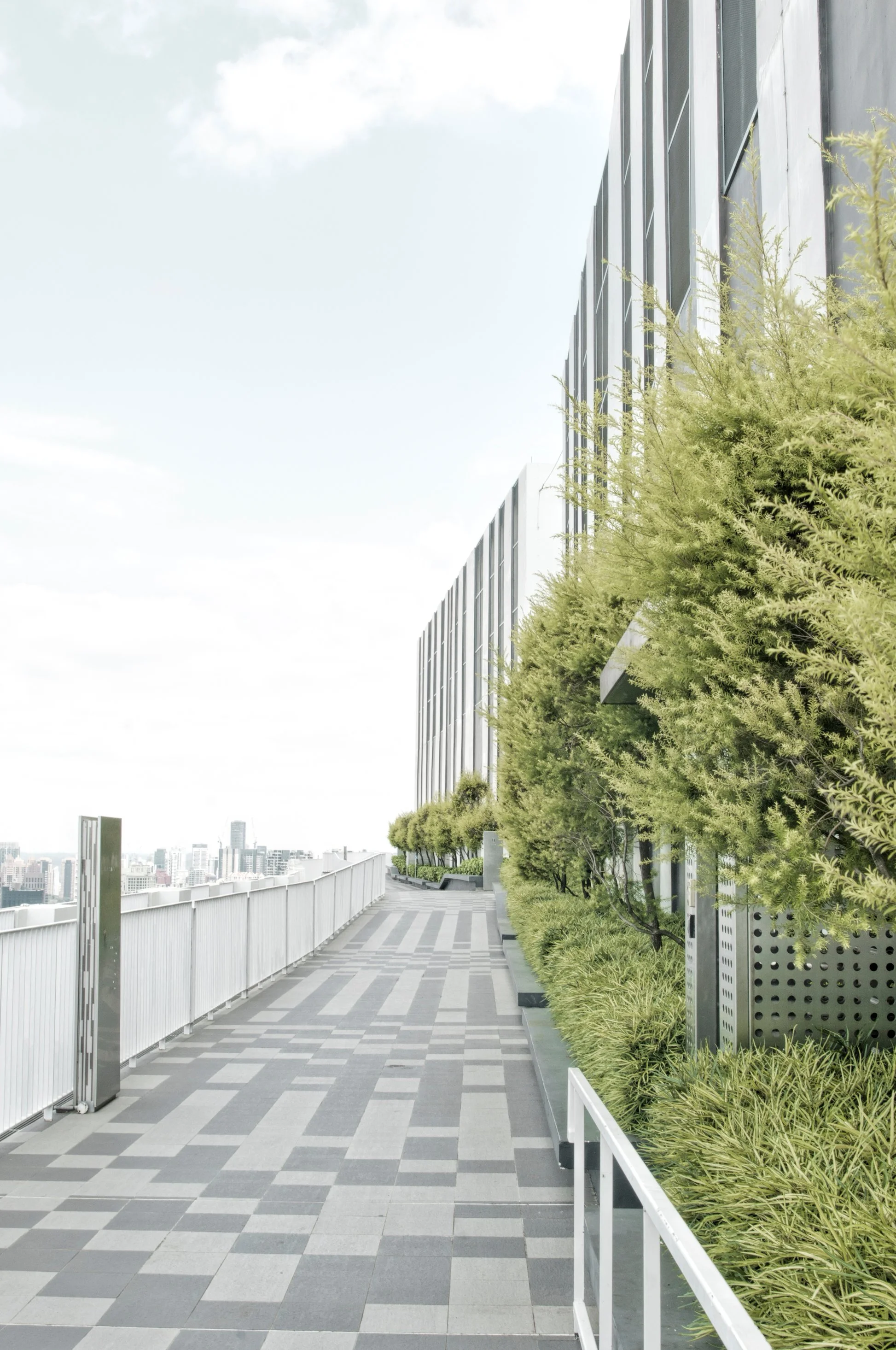 A modern outdoor terrace with a checkered gray and beige tile pattern, lined with lush green plants in planters along a metal railing, with a city skyline visible in the distance under a partly cloudy sky.