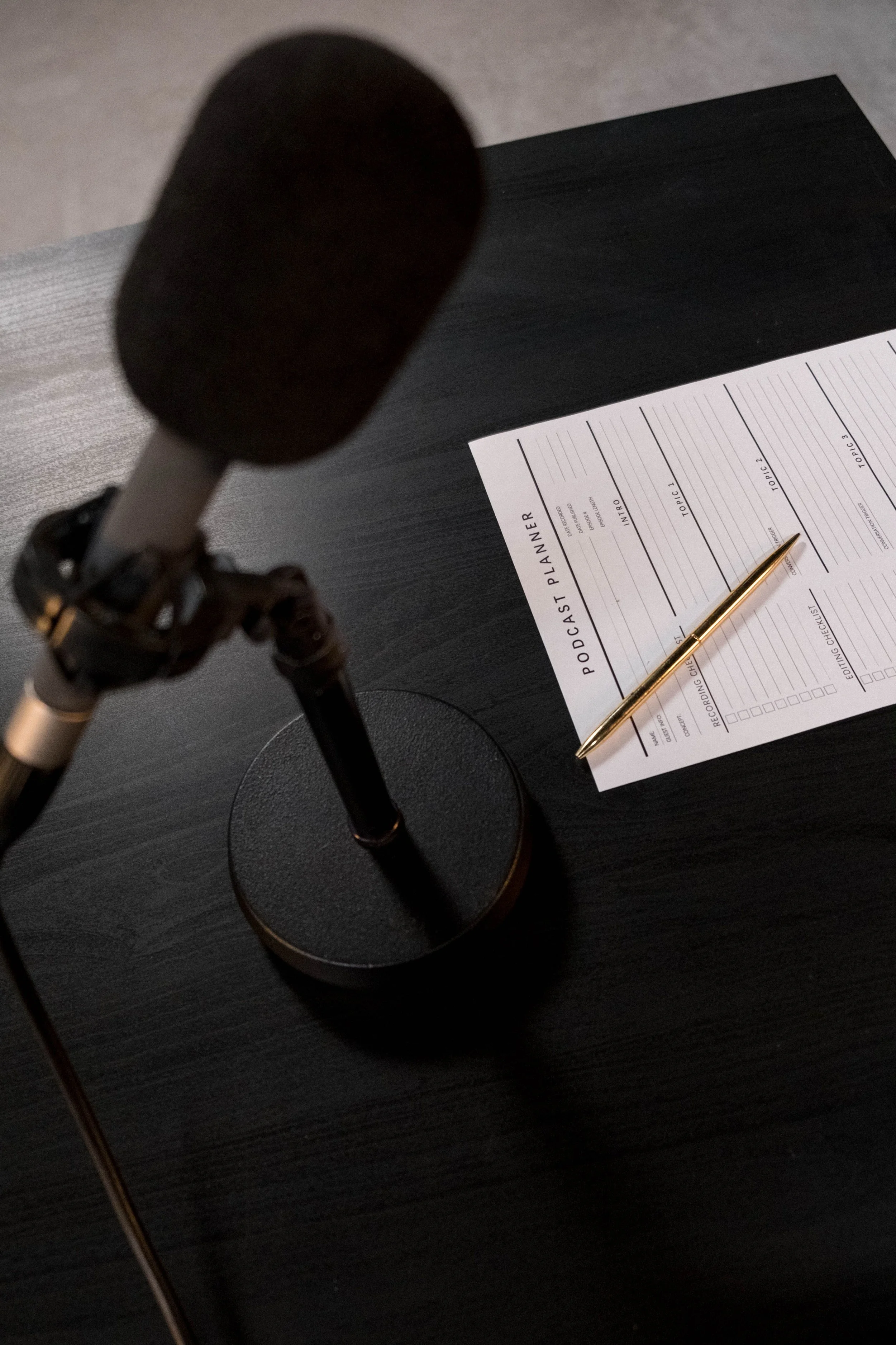 Microphone, ordinateur portable noir, planificateur de podcast avec un stylo doré dessus, sur une table en bois foncé.