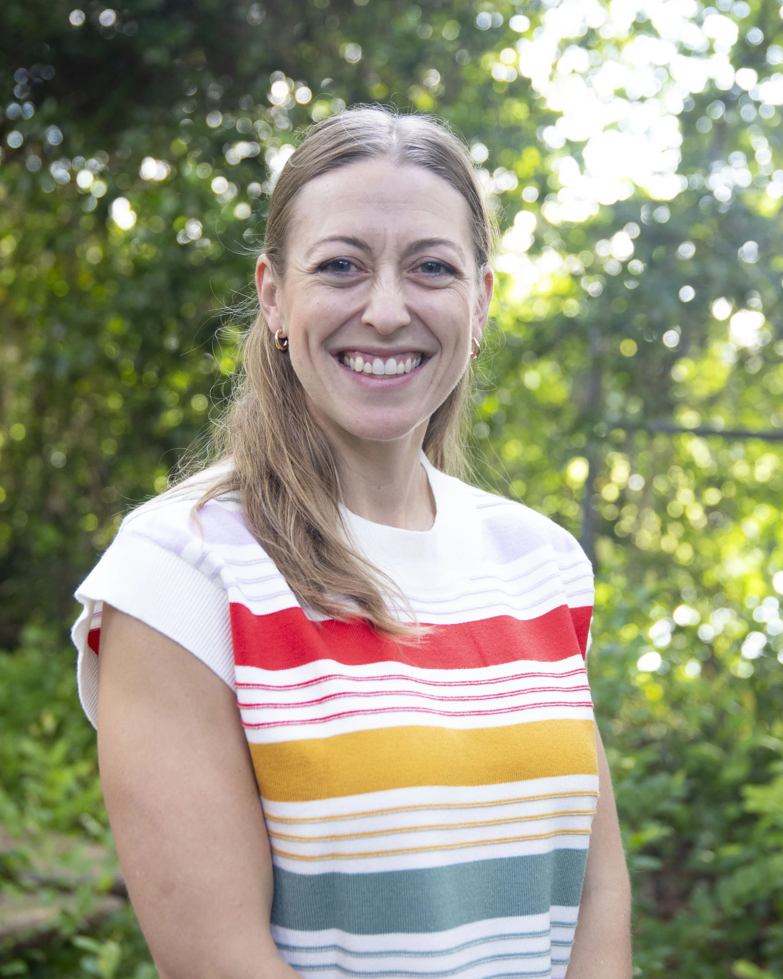 A smiling woman with long light brown hair and blue eyes, wearing a colorful striped sweater, standing outdoors with a blurred green foliage background.