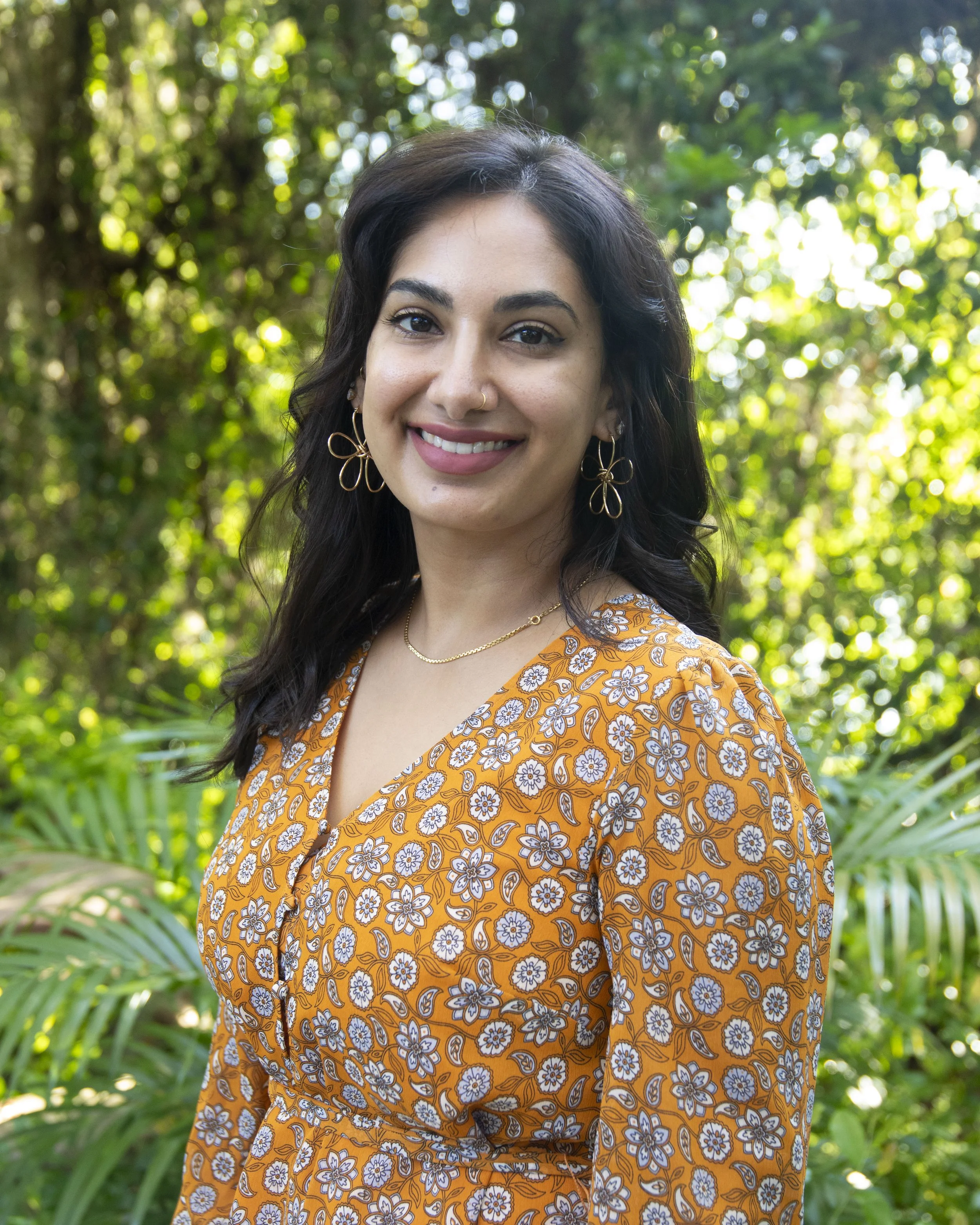 A woman outdoors in a lush, green setting with trees and plants, smiling at the camera. She is wearing a floral patterned orange dress, gold jewelry including earrings and a necklace, and has dark, wavy hair.
