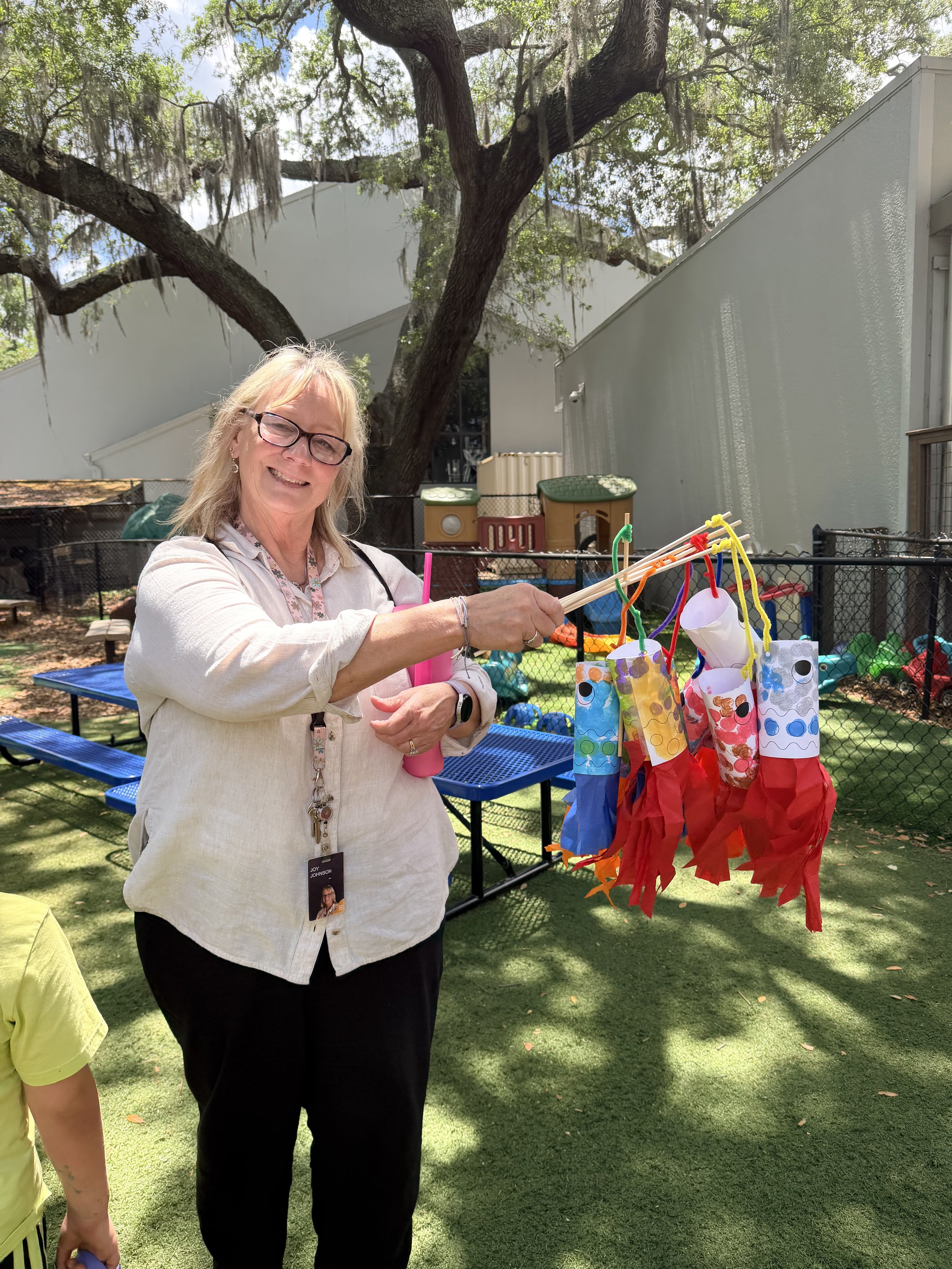 A woman smiling and holding colorful paper lanterns in an outdoor setting with large trees, a building, and a fence in the background.