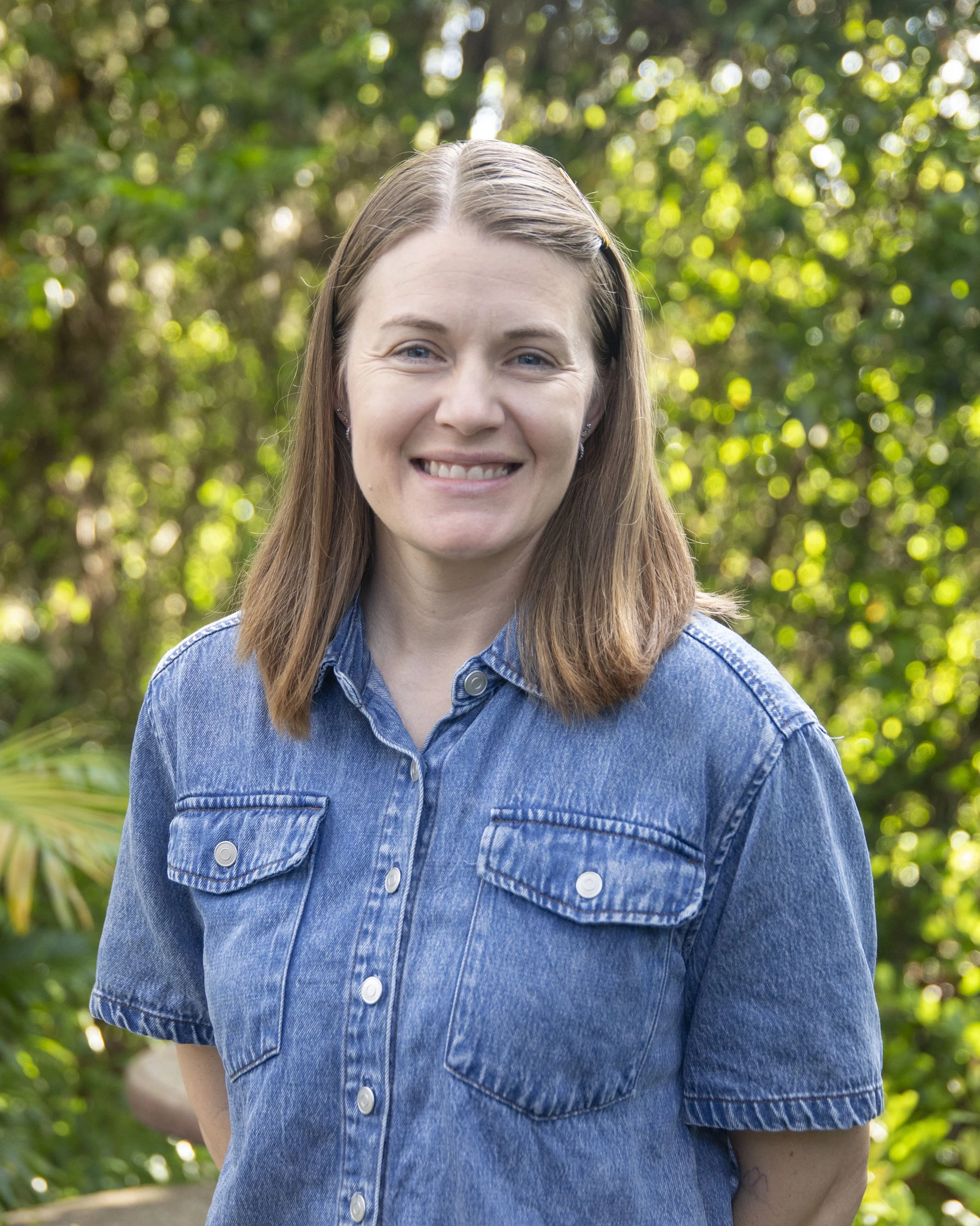 Close-up of a woman with brown hair wearing a denim shirt, smiling outdoors with green foliage in the background.