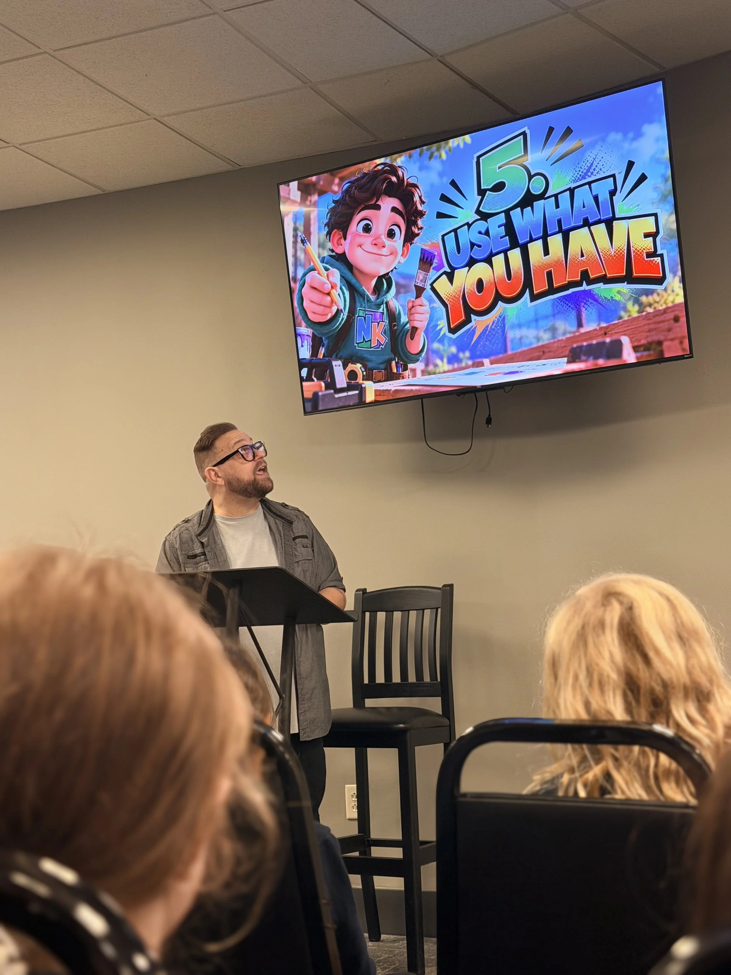 A man with glasses standing in front of a microphone in a room with beige walls and gray ceiling tiles, facing an audience with only the tops of their heads visible, while a large colorful screen above displays a cartoon boy with brown hair and a green shirt, holding a paintbrush, along with the text '5. USE WHAT YOU HAVE'.