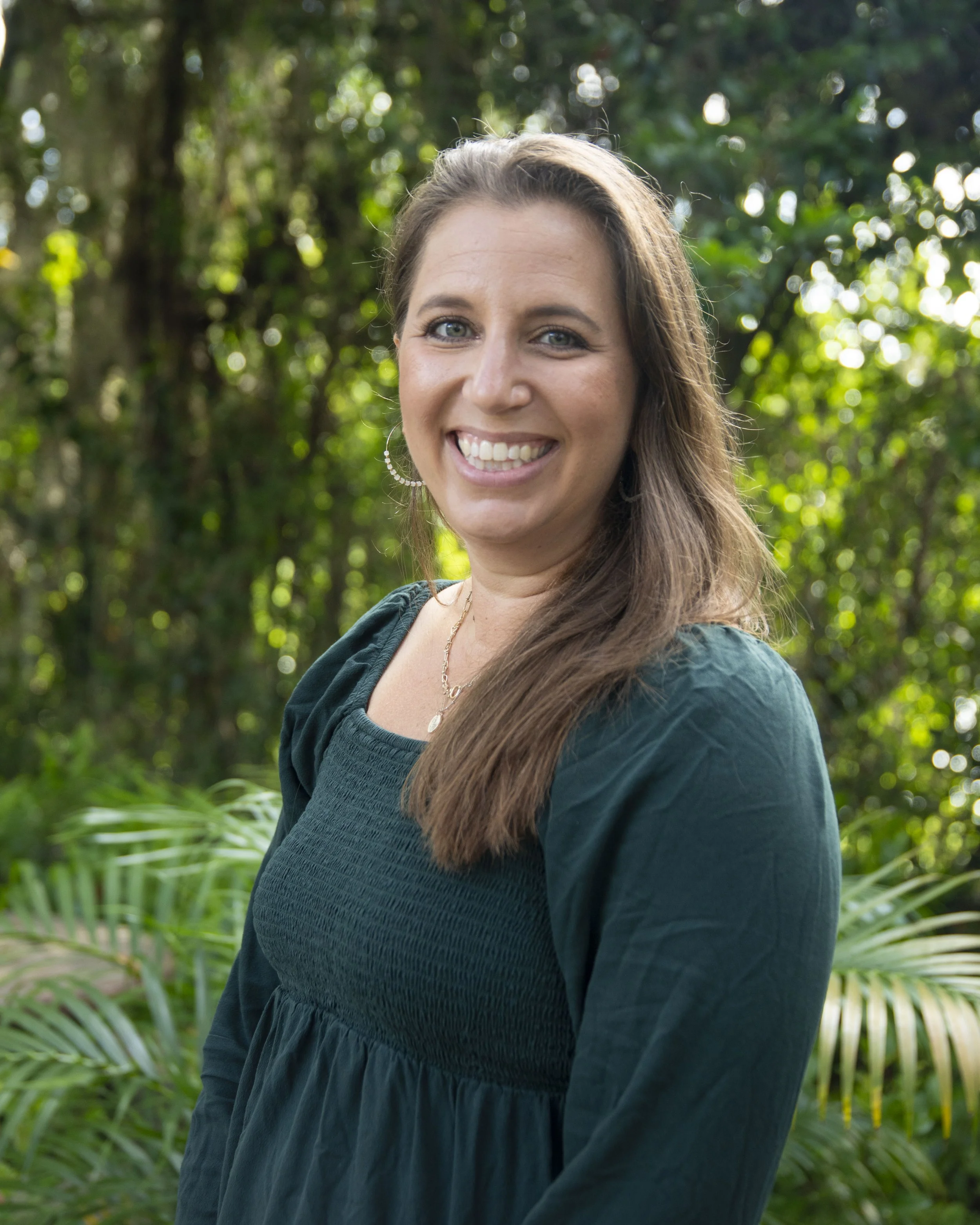 A smiling woman with light brown hair and blue eyes standing outdoors with greenery in the background.