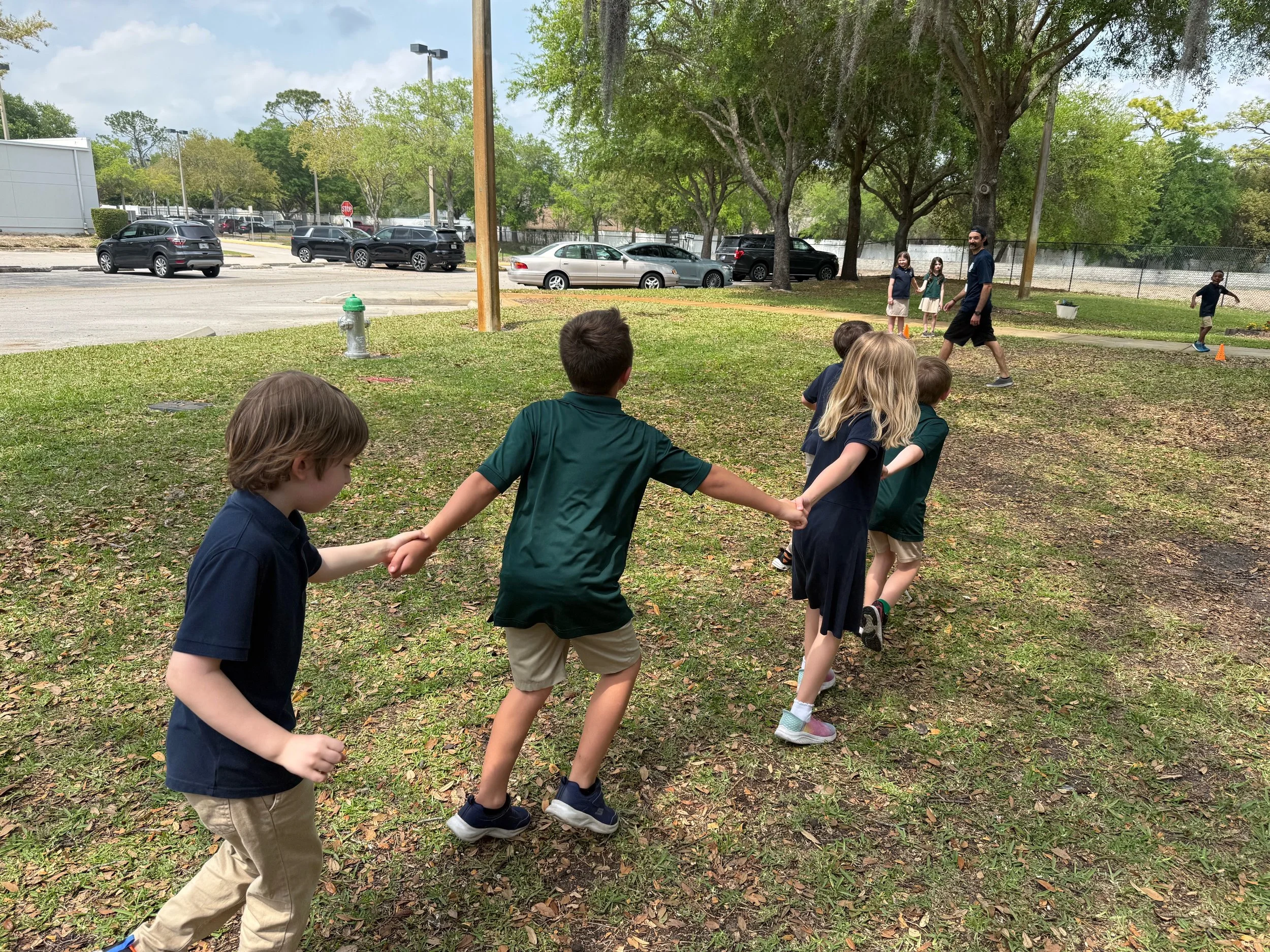 Children holding hands in a line outdoors with trees, a fire hydrant, and parked cars in the background.